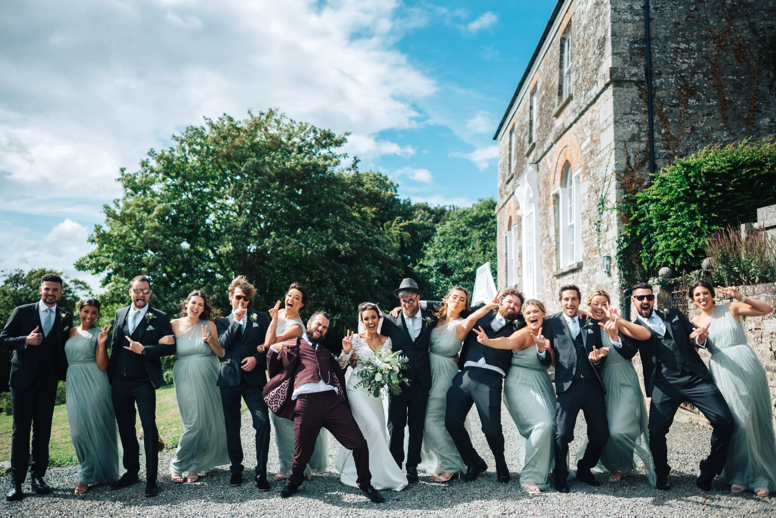 Group of people dressed in wedding attire posing outdoors with a stone building and trees in the background.