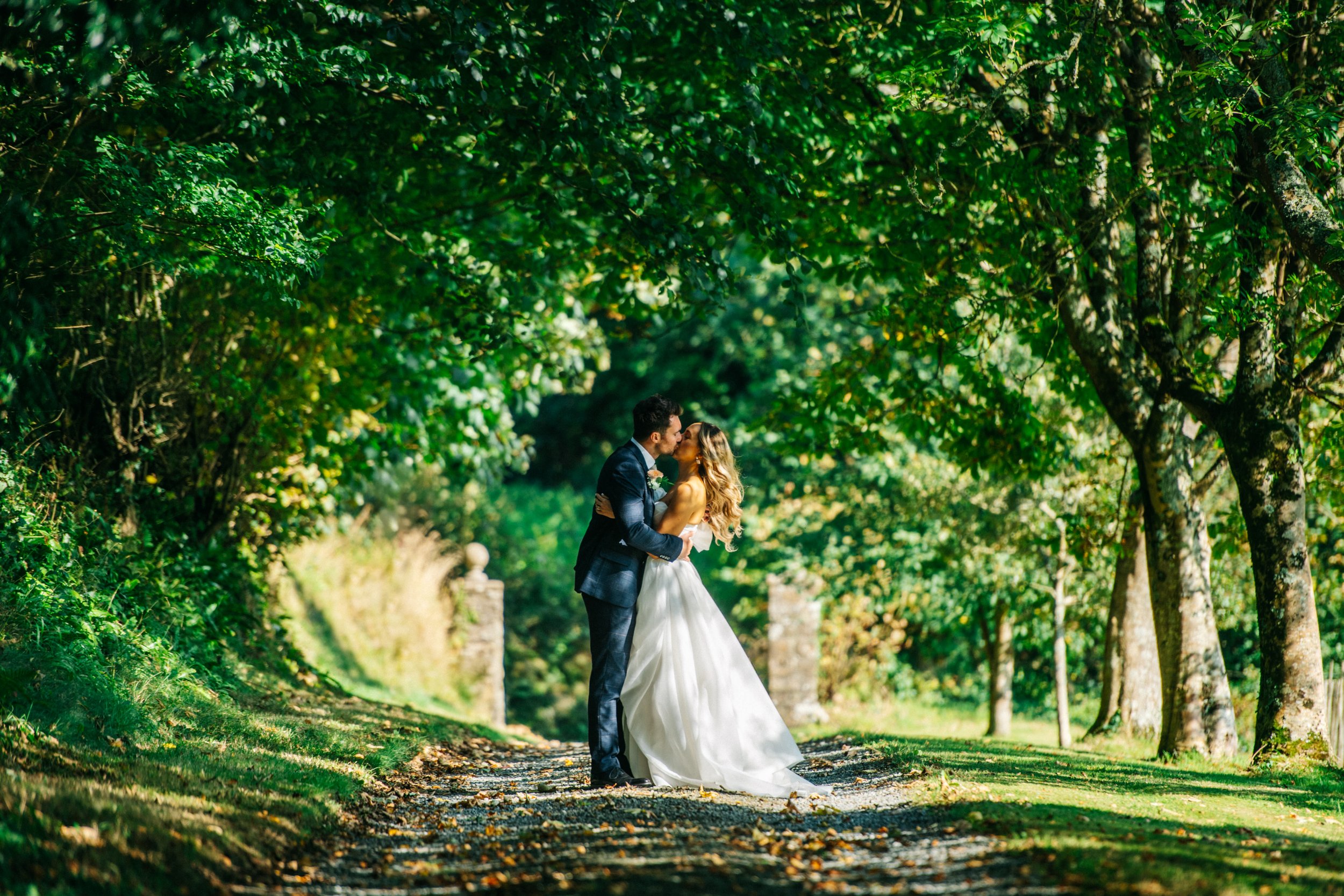 A newlywed couple in wedding attire sharing a kiss on a tree-lined path in a park.