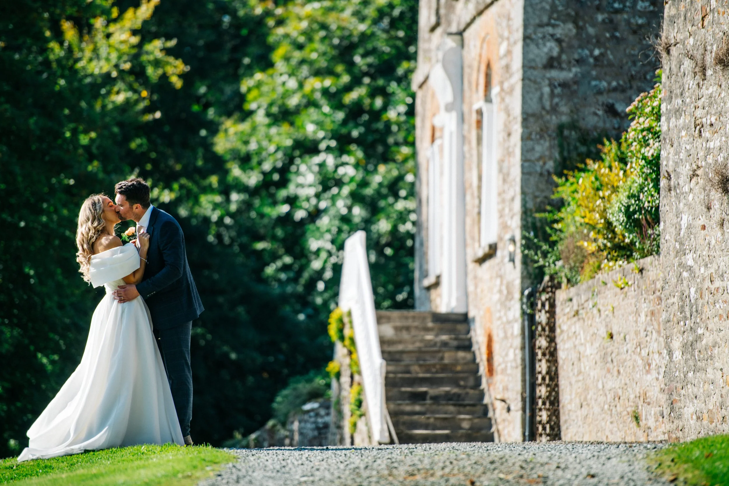 A bride and groom are kissing outside a stone building on a sunny day.
