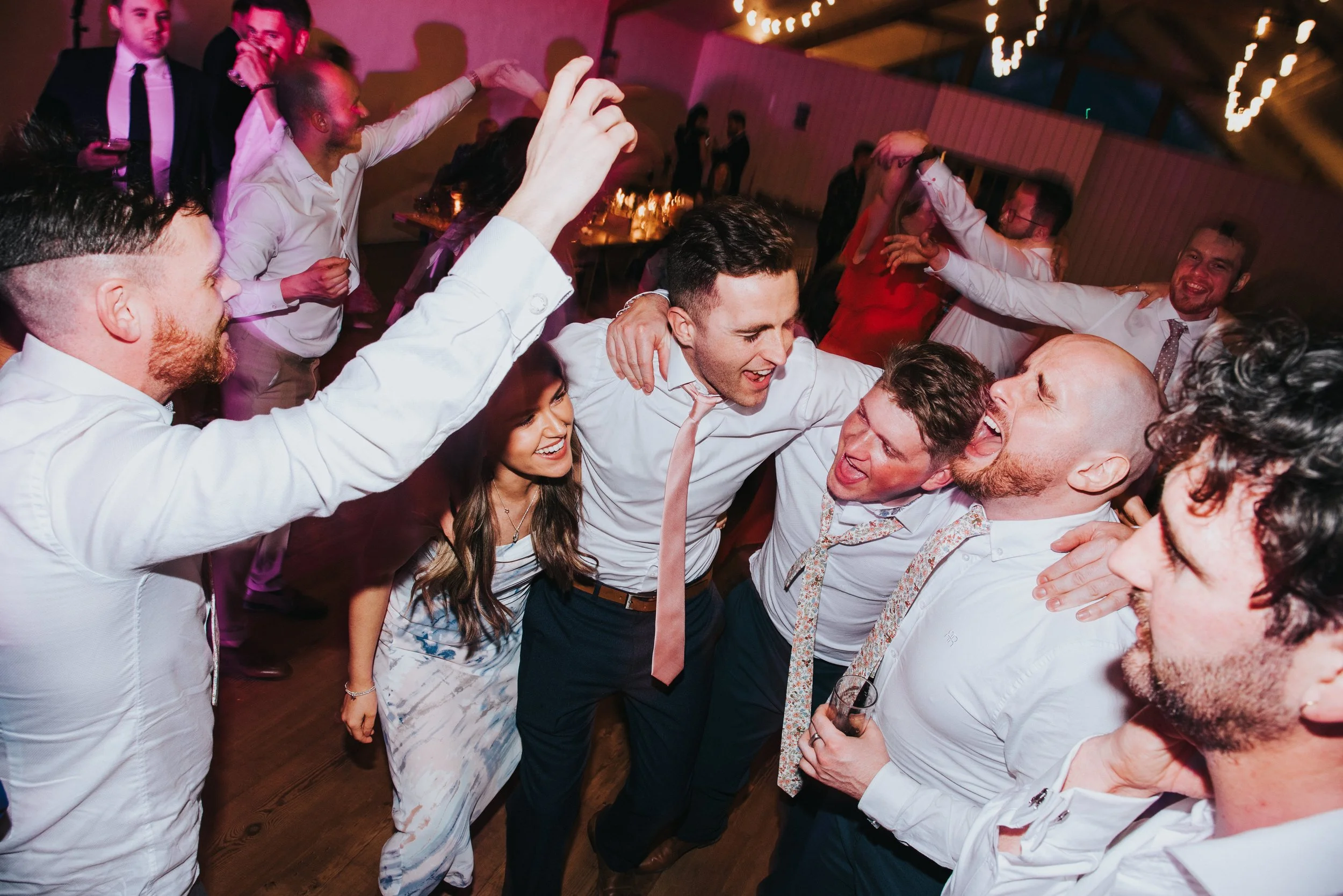 People dancing and celebrating at a party, smiling, embracing, and having fun in a decorated room with string lights.