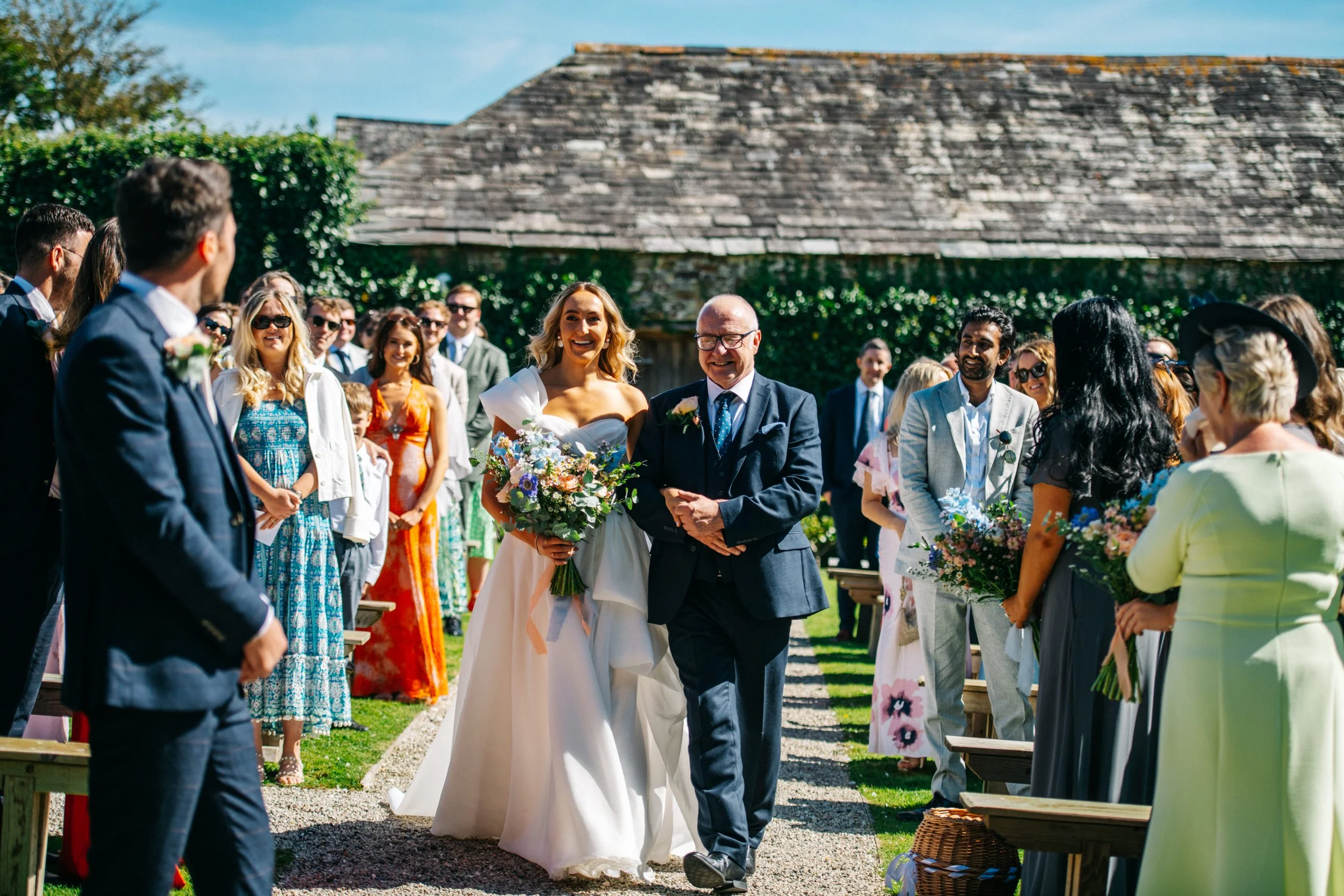 A bride in a white wedding gown walking down the aisle with an older man, likely her father, at an outdoor wedding ceremony, with guests standing and smiling.