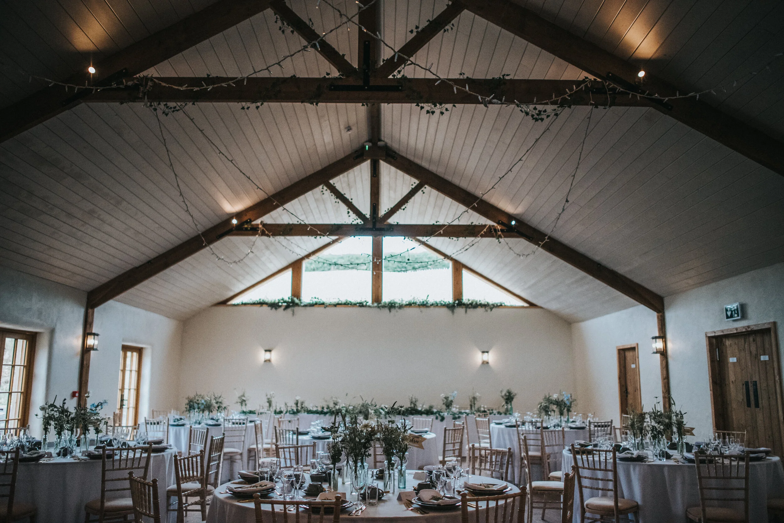 Decorated event hall with round tables, chairs, and floral centerpieces, under a high ceiling with string lights and wooden beams.