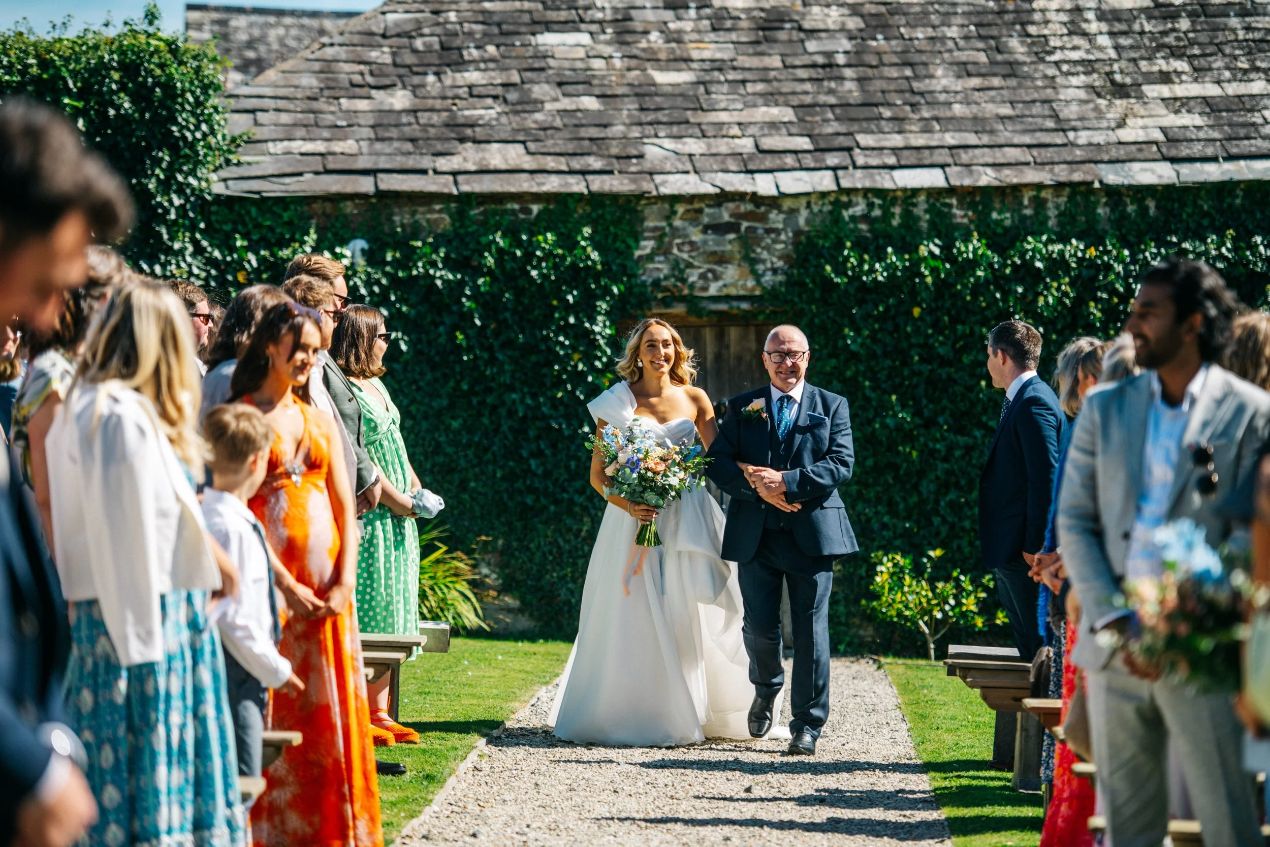 A bride walking down the aisle in a garden, accompanied by a man, possibly her father, during a wedding ceremony with guests seated on either side.