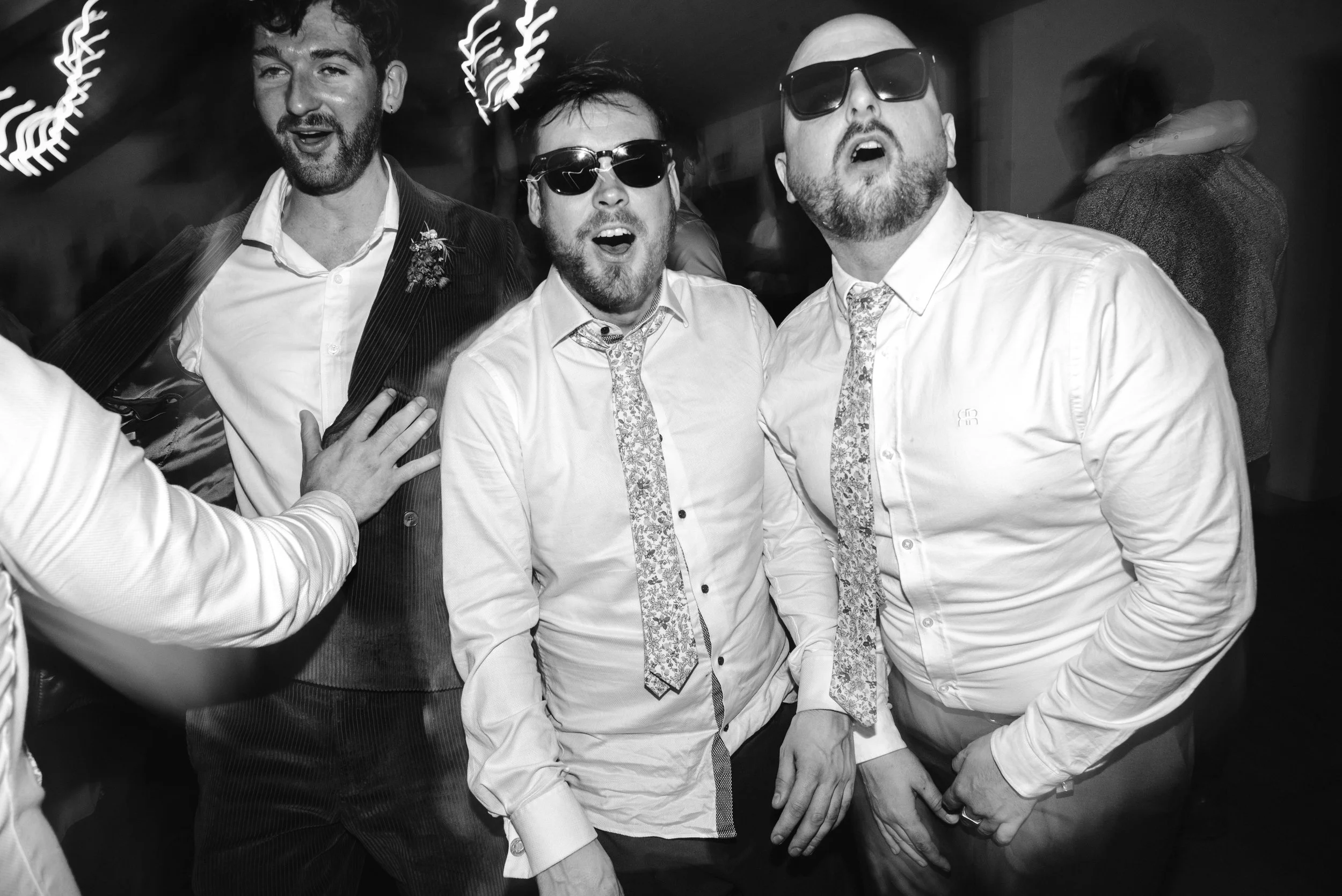 Three men wearing white shirts, patterned ties, and sunglasses are at a social gathering, with one man in the background wearing a dark jacket.