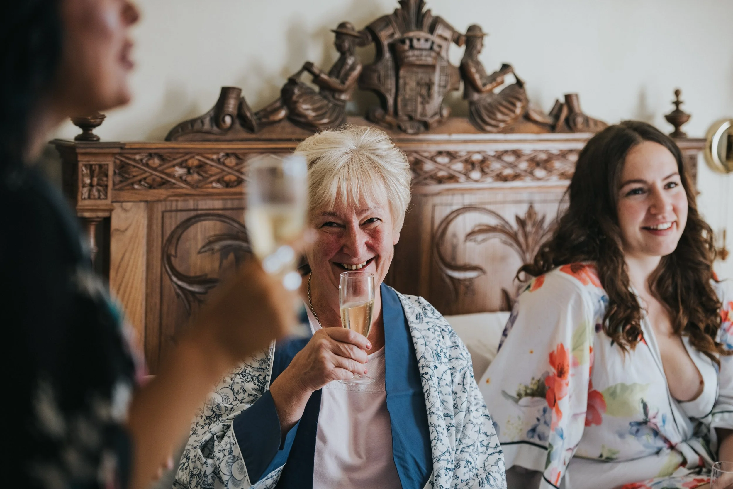 People celebrating with glasses of champagne, woman in the middle smiling and holding a glass of champagne, woman to her right smiling, woman on the left blurred holding a glass of champagne, sitting at a wooden table with detailed carved woodwork in