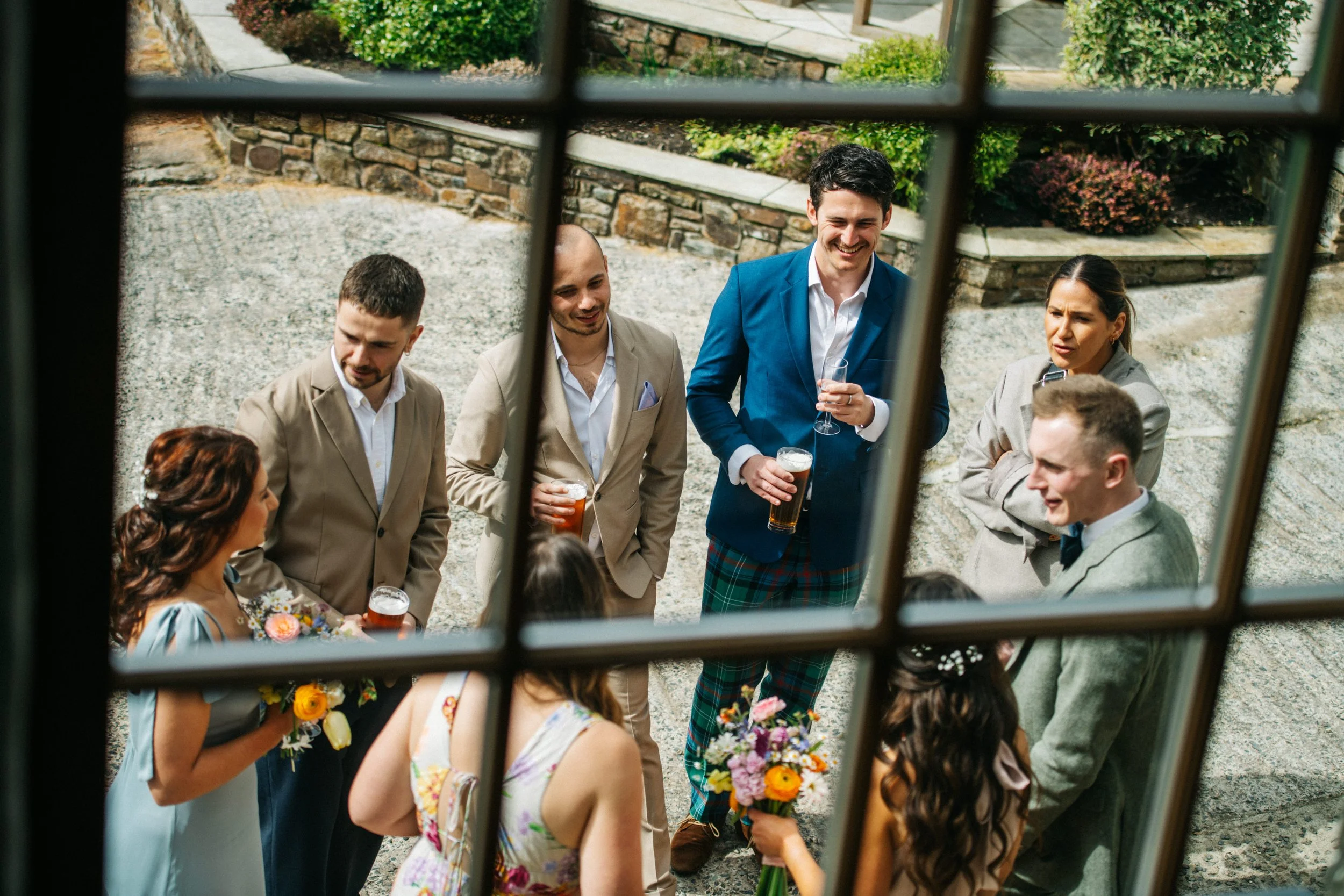 Group of people socializing outside, seen through a window with black grid muntins. They are holding drinks and flowers, dressed in formal attire, engaged in conversation, smiling, and enjoying a sunny day in a garden setting.