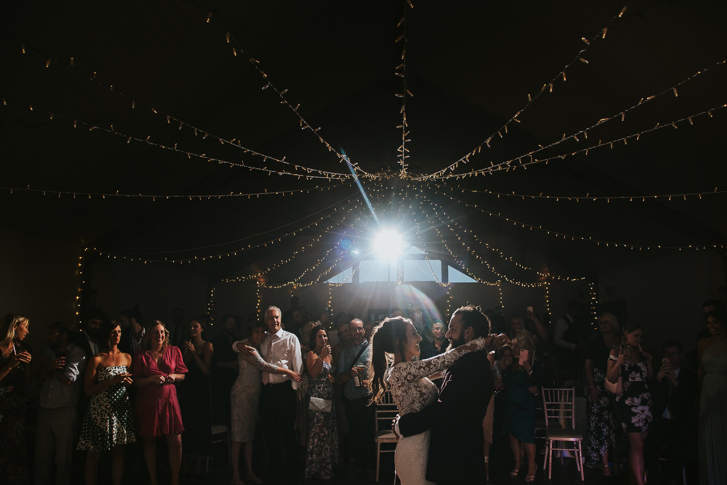 Couple dancing at their wedding reception under string lights, with guests watching and taking photos in the background.
