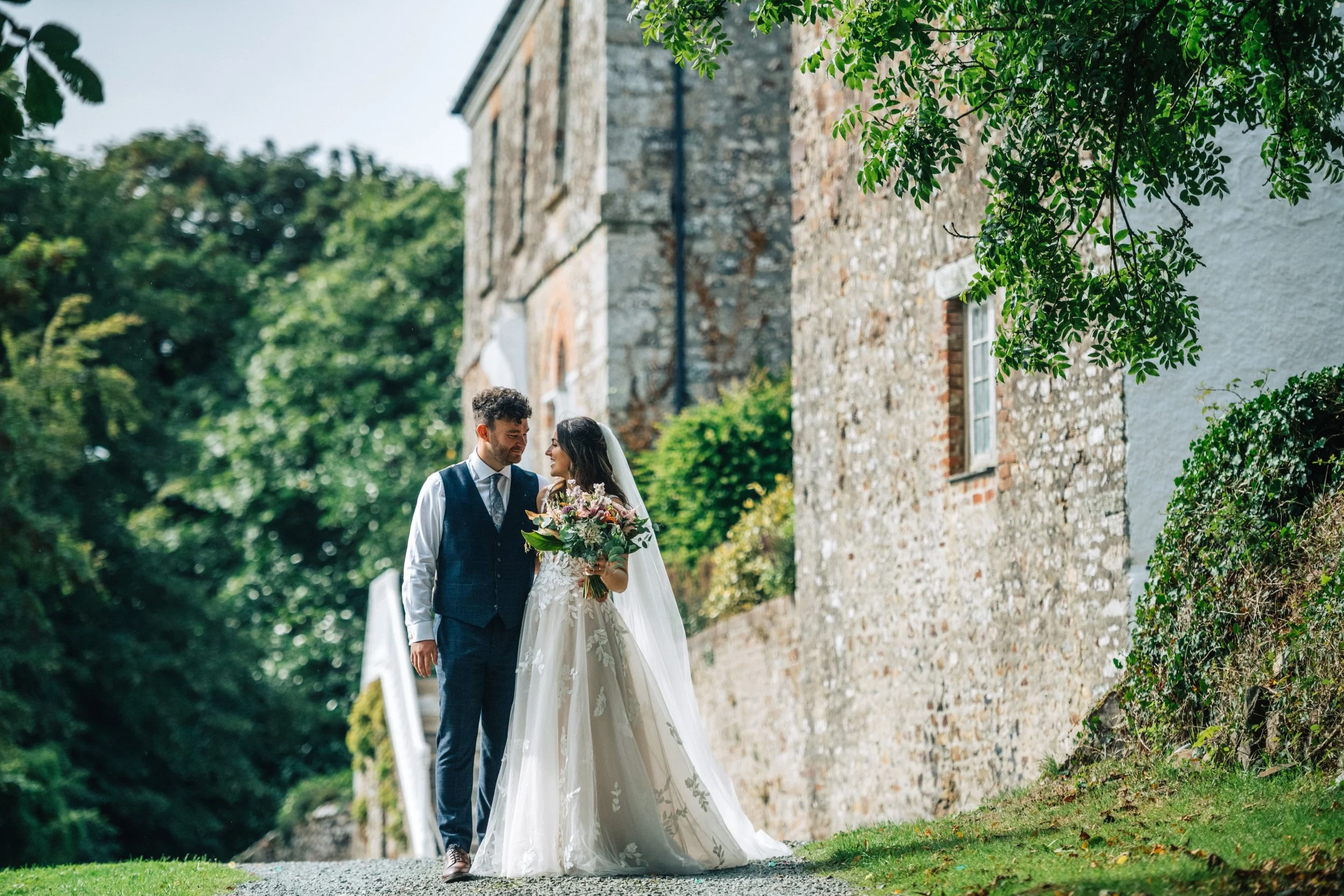 A bride and groom standing outdoors on a cobblestone path with a stone building in the background, surrounded by greenery.