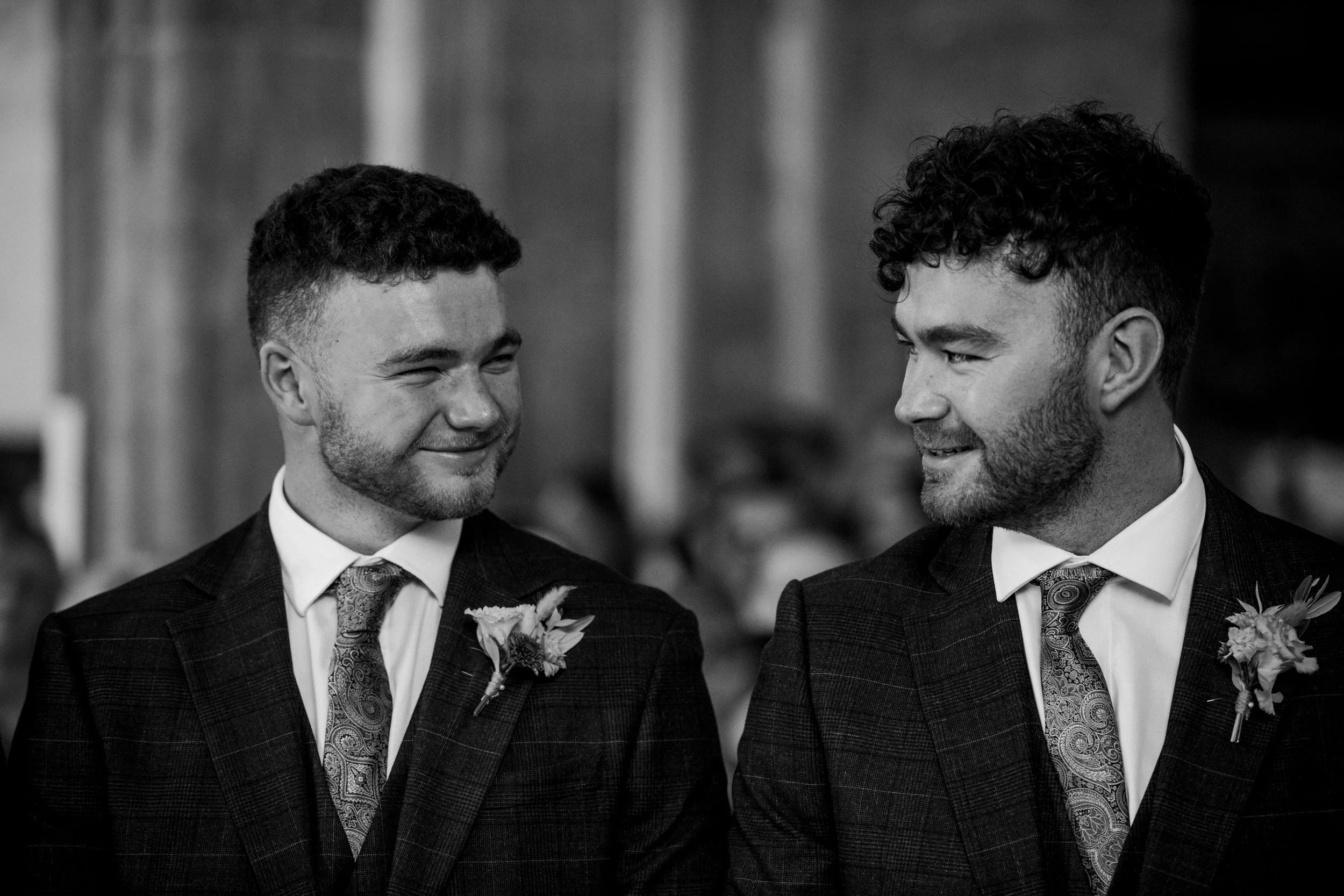 Two men wearing suits with boutonnieres, smiling at each other in a formal indoor setting.