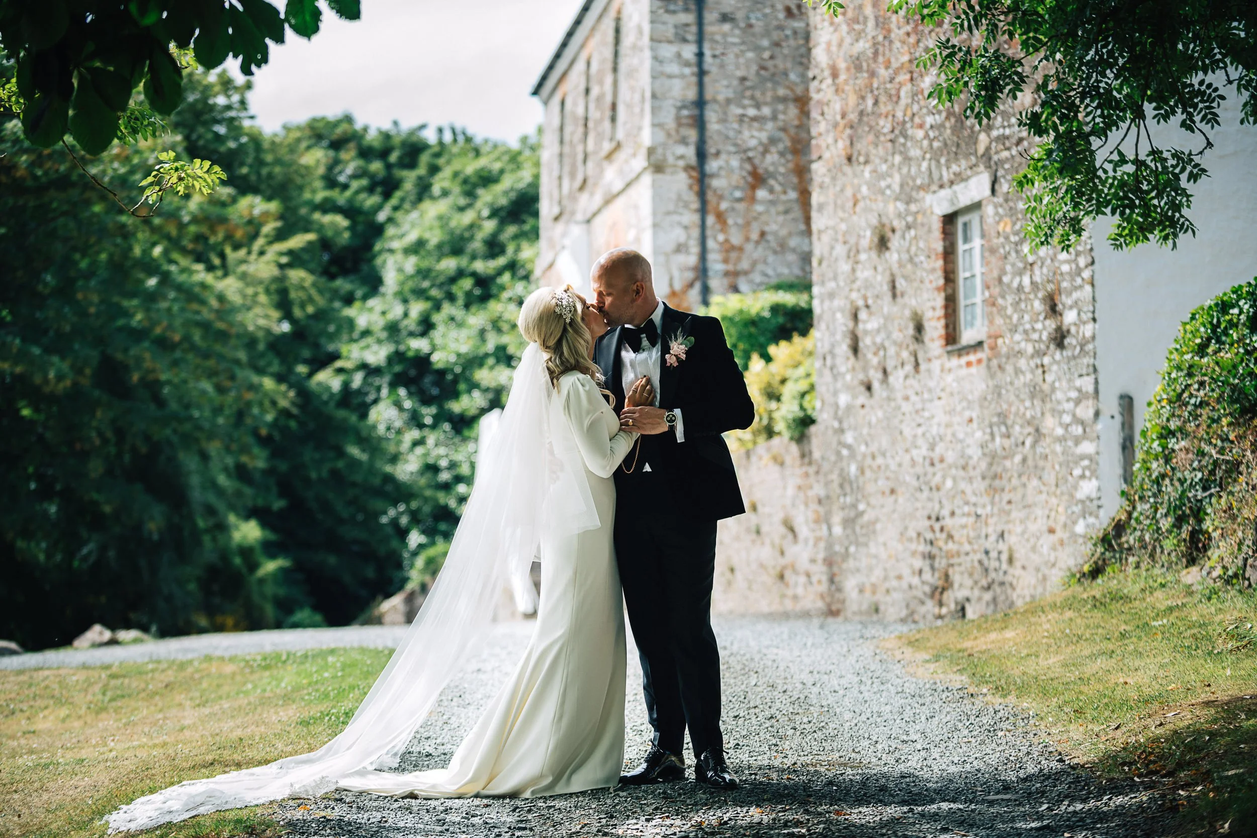 A bride and groom share a kiss outdoors on a gravel path with greenery and a stone building in the background.