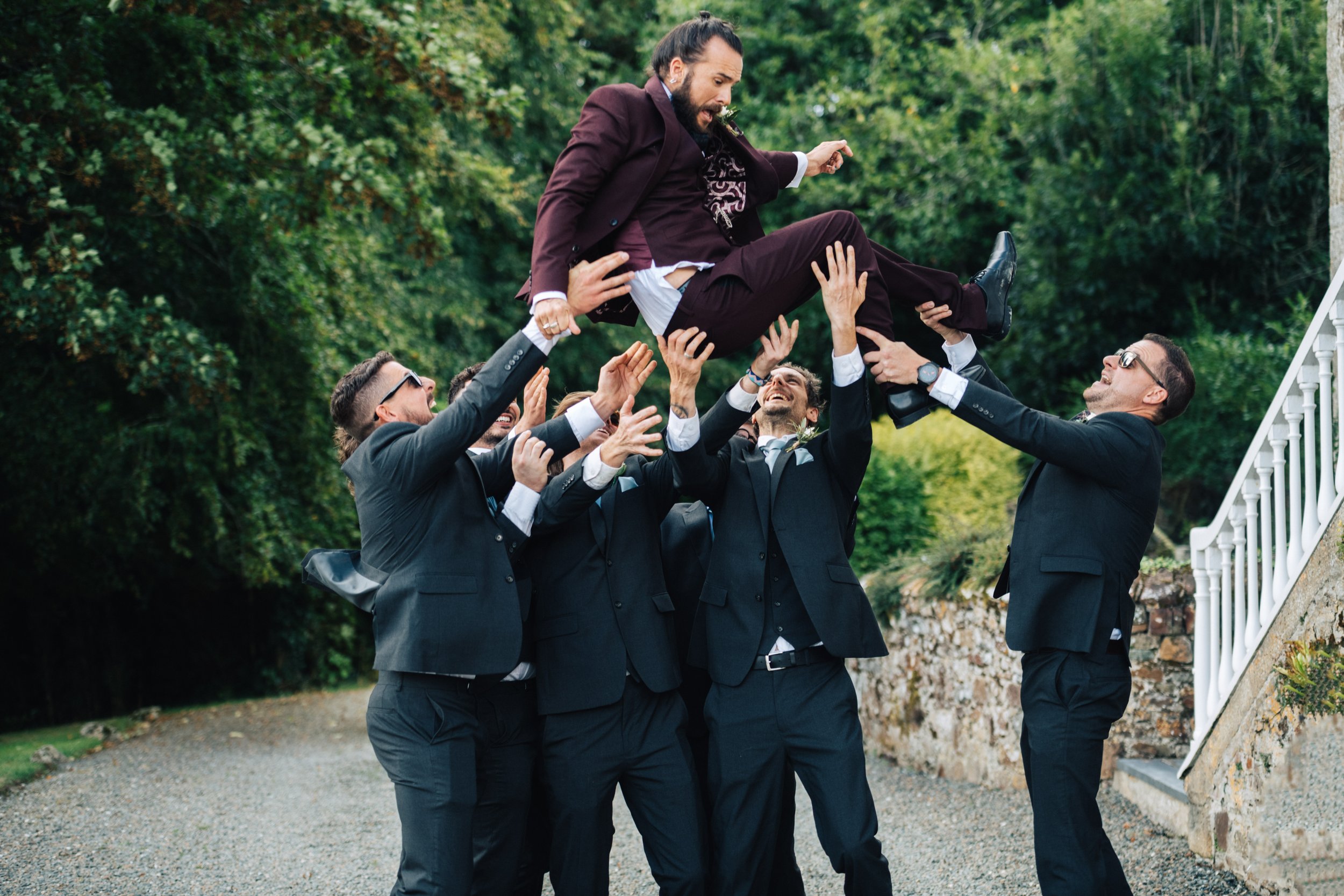 A group of men dressed in suits lifting a man in a maroon suit in the air outdoors in front of green trees.