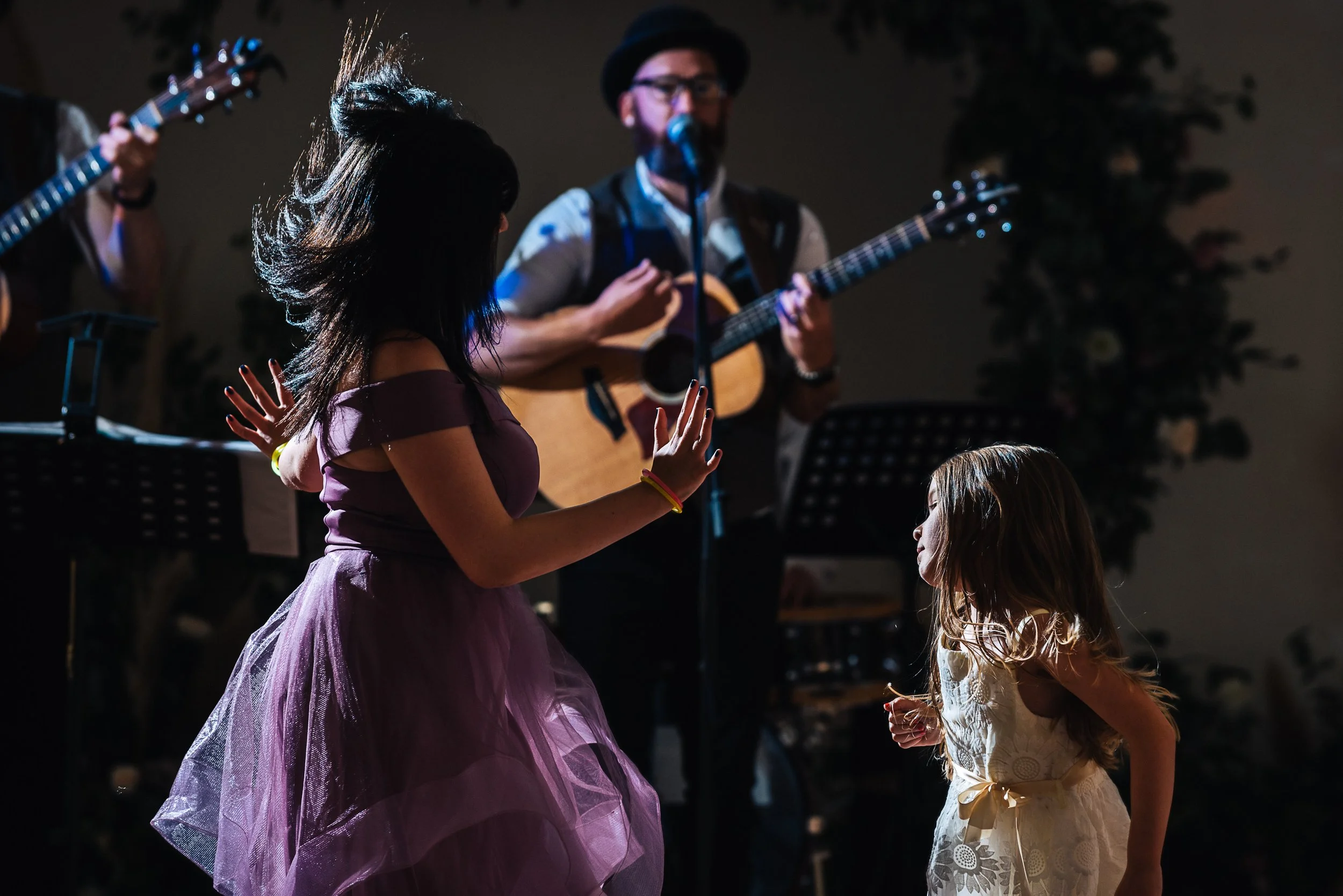 Two young girls dancing in front of a musician playing guitar and singing into a microphone at an indoor event with a musical band.