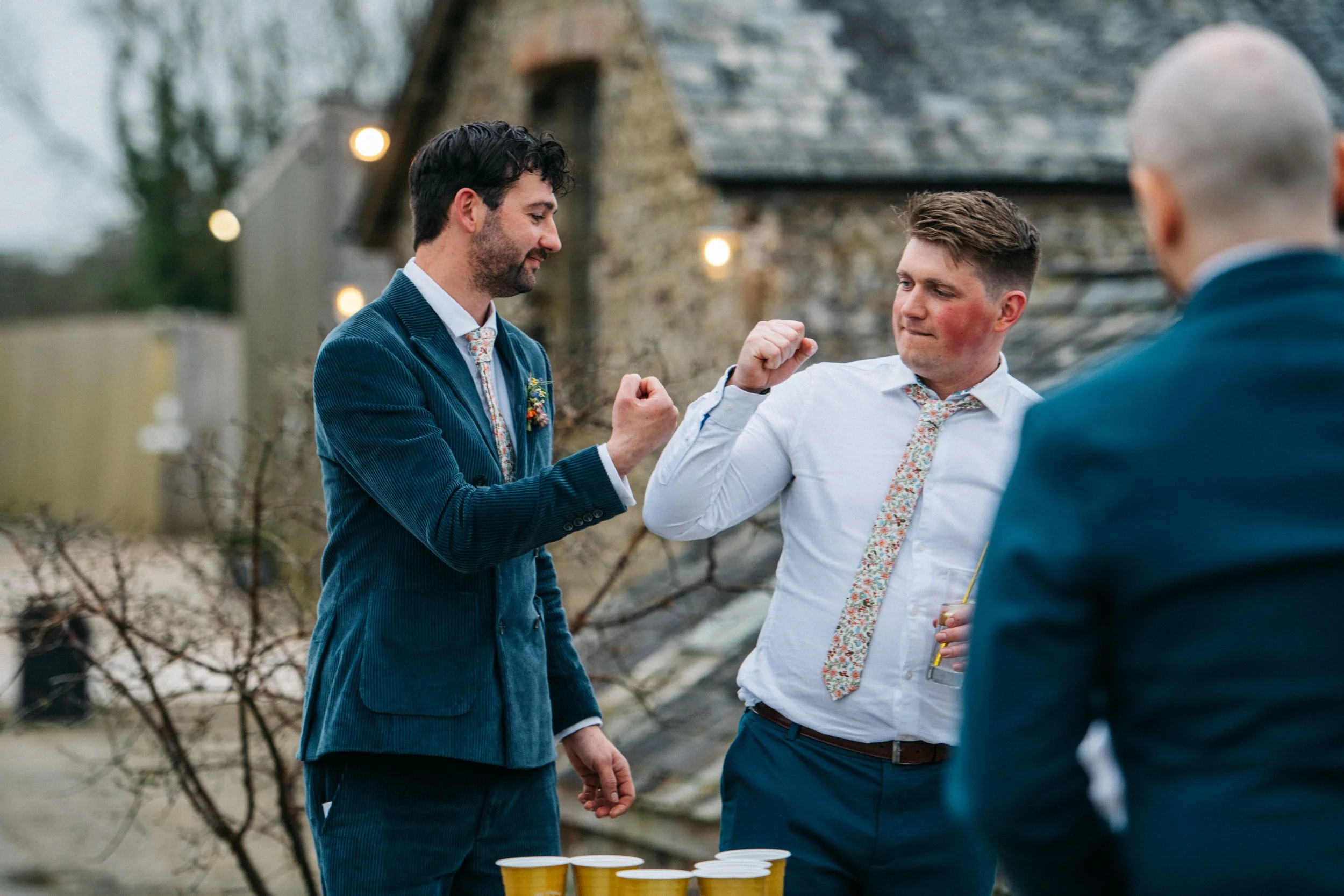 Three men in suits at an outdoor gathering, two of them are playfully flexing their arms, with a table of yellow cups in front of them, and a rustic building with string lights in the background.