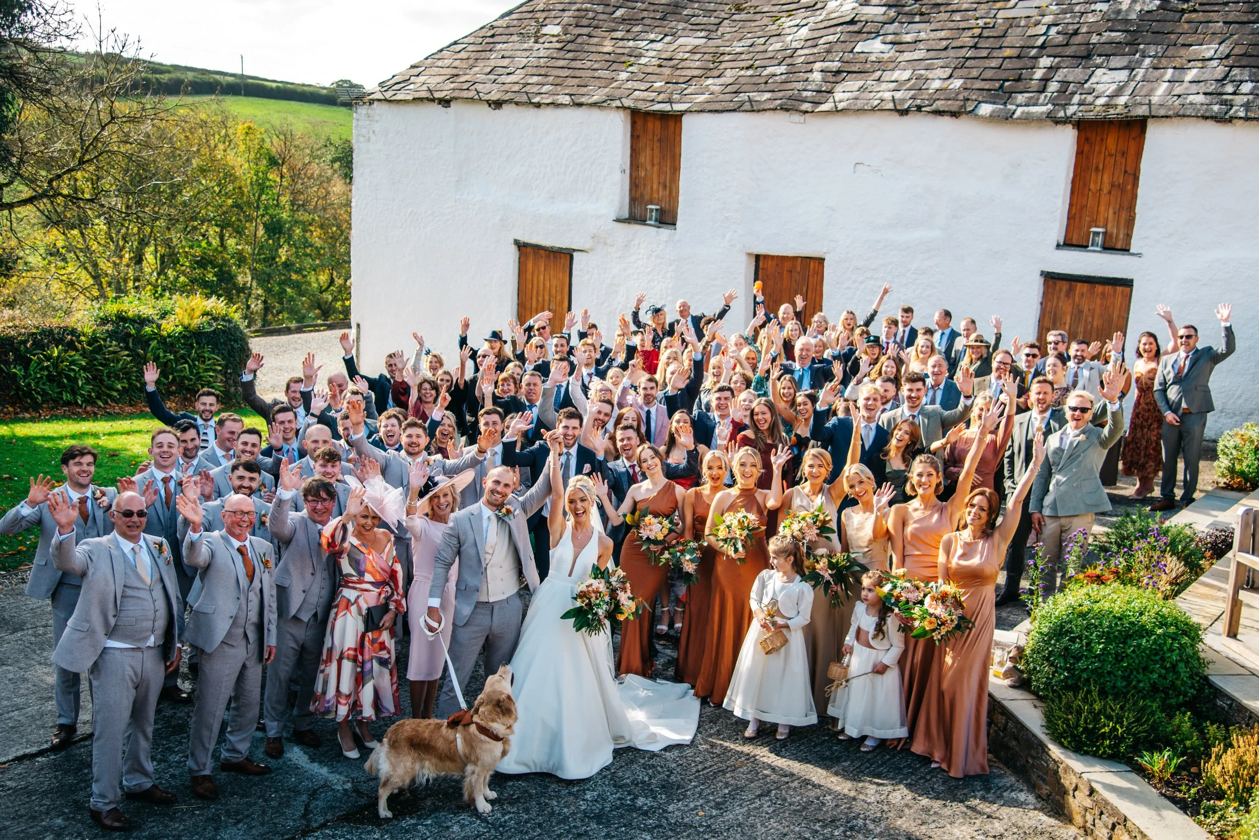 A large group of wedding guests and the wedding couple gathered outdoors in front of a white building, all waving at the camera on a sunny day, with some holding bouquets and others wearing formal suits and dresses.