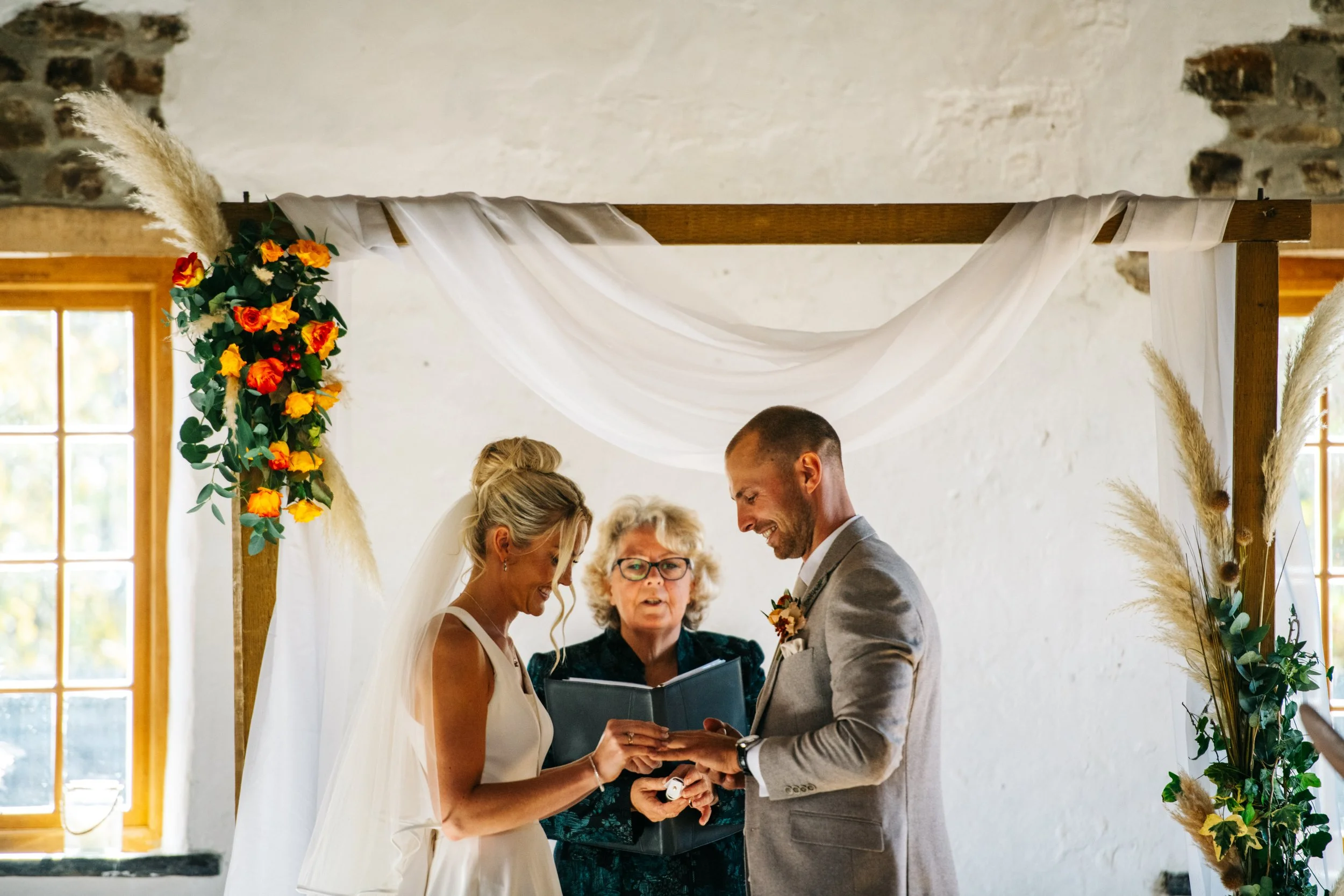 A bride and groom exchanging rings during a wedding ceremony, officiated by a woman standing between them, in a decorated indoor setting with windows and white walls.