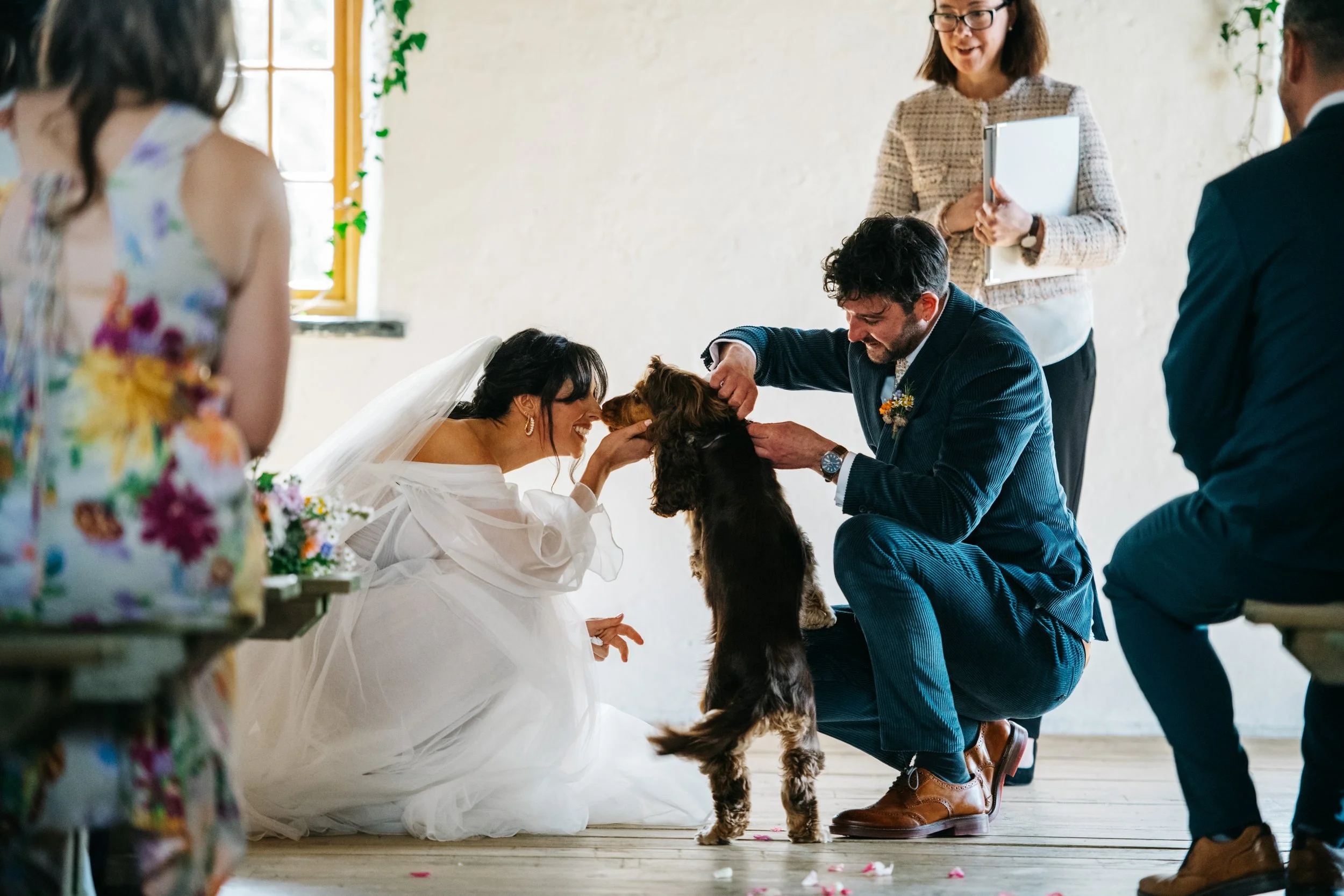 A bride and groom at a wedding ceremony with a dog. The bride, in a white wedding dress and veil, touches the dog's face. The groom, in a dark suit, holds and interacts with the dog. A woman standing behind them holds a book, observing the moment. Gu