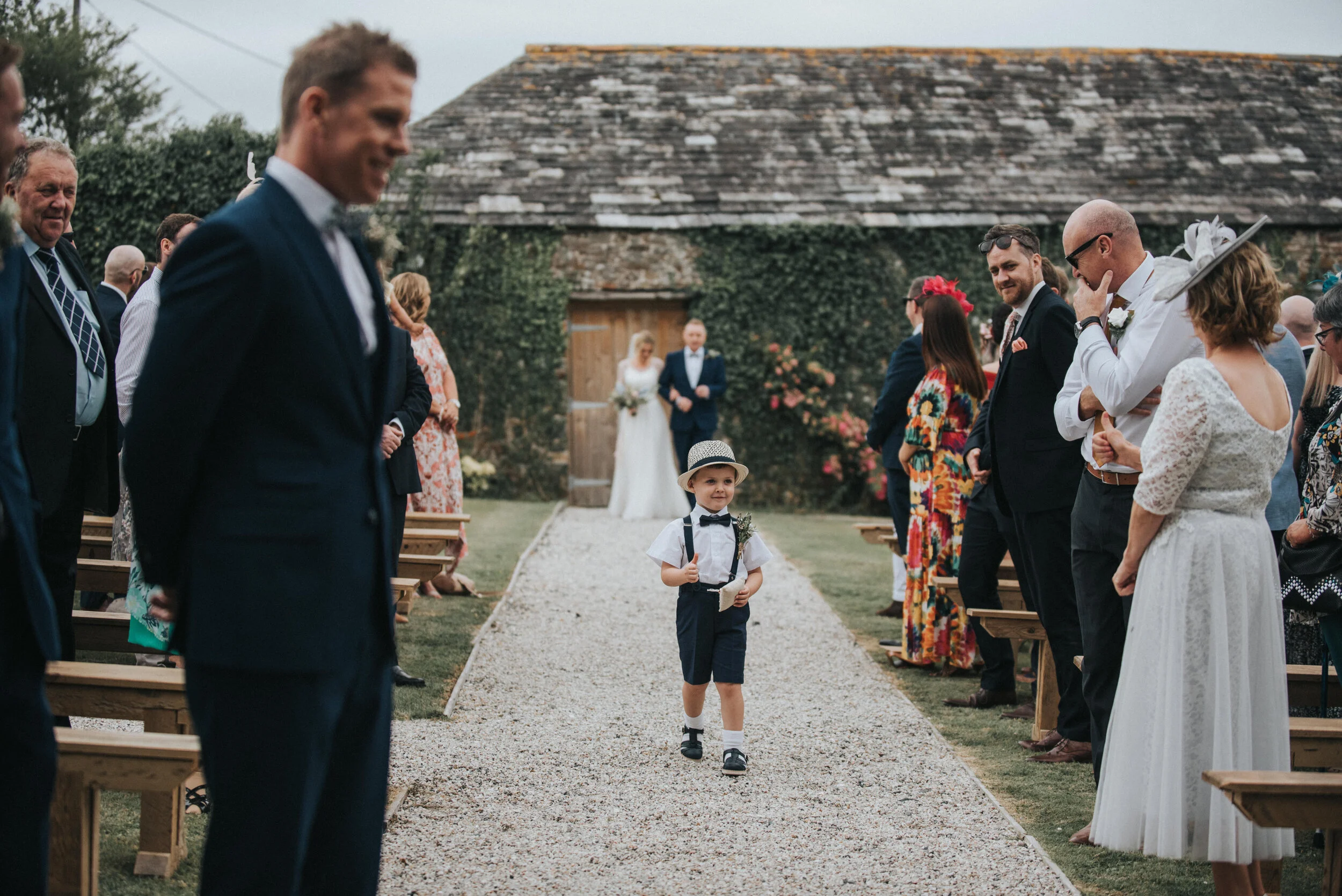 A young boy dressed in a white shirt, navy shorts, suspenders, and a bow tie walks down the aisle at an outdoor wedding, surrounded by seated guests and a couple of brides and grooms in the background
