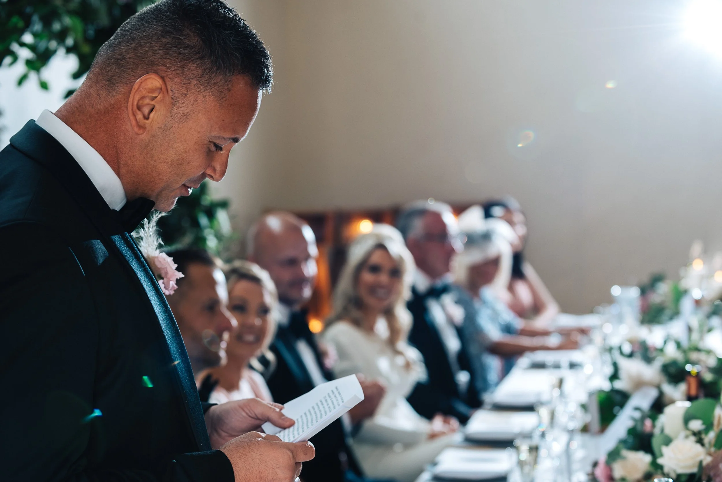 A man in formal attire reading from a paper at a wedding reception table filled with guests, flowers, and decorations.