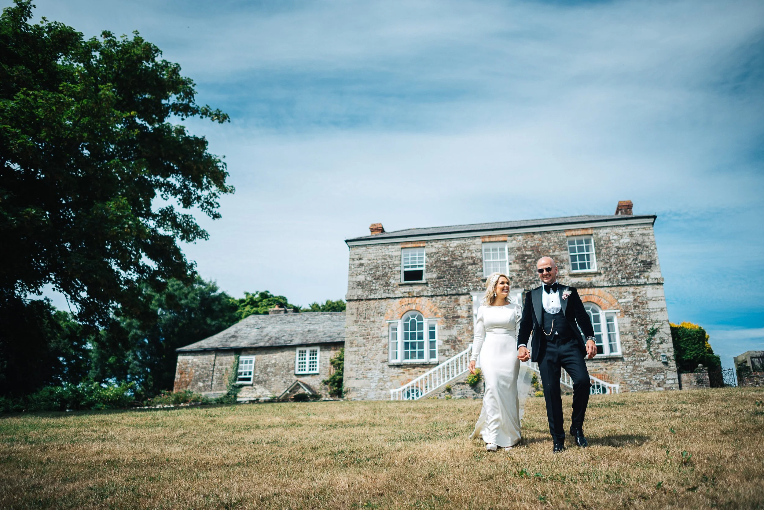 Bride and groom holding hands and walking outside a historic brick house with large windows on a sunny day.