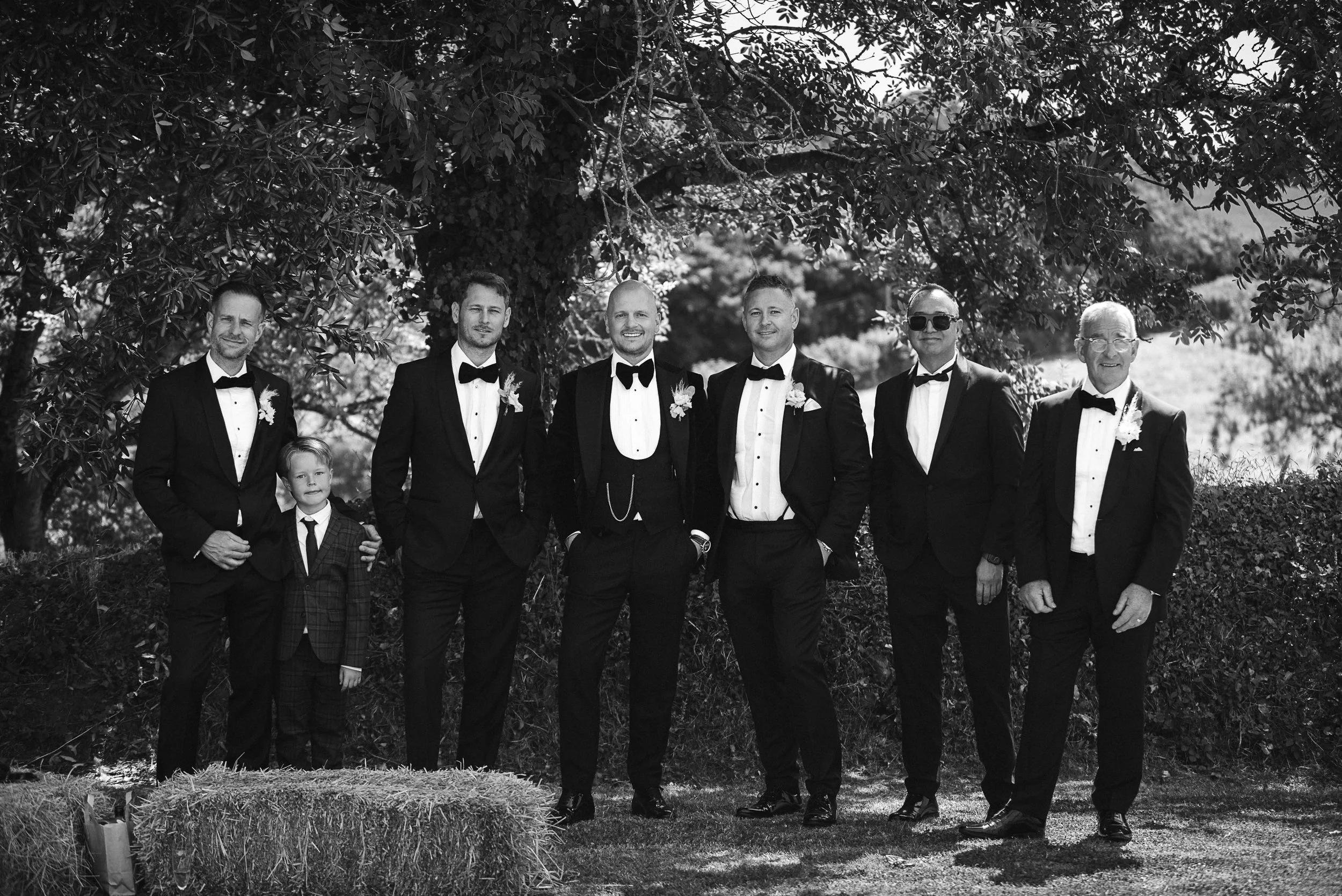 A group of seven men, dressed in tuxedos, standing outdoors under a large tree. One young boy in a suit is in front of the group. They are all smiling, with some wearing sunglasses and boutonnières, suggesting a wedding or formal event.