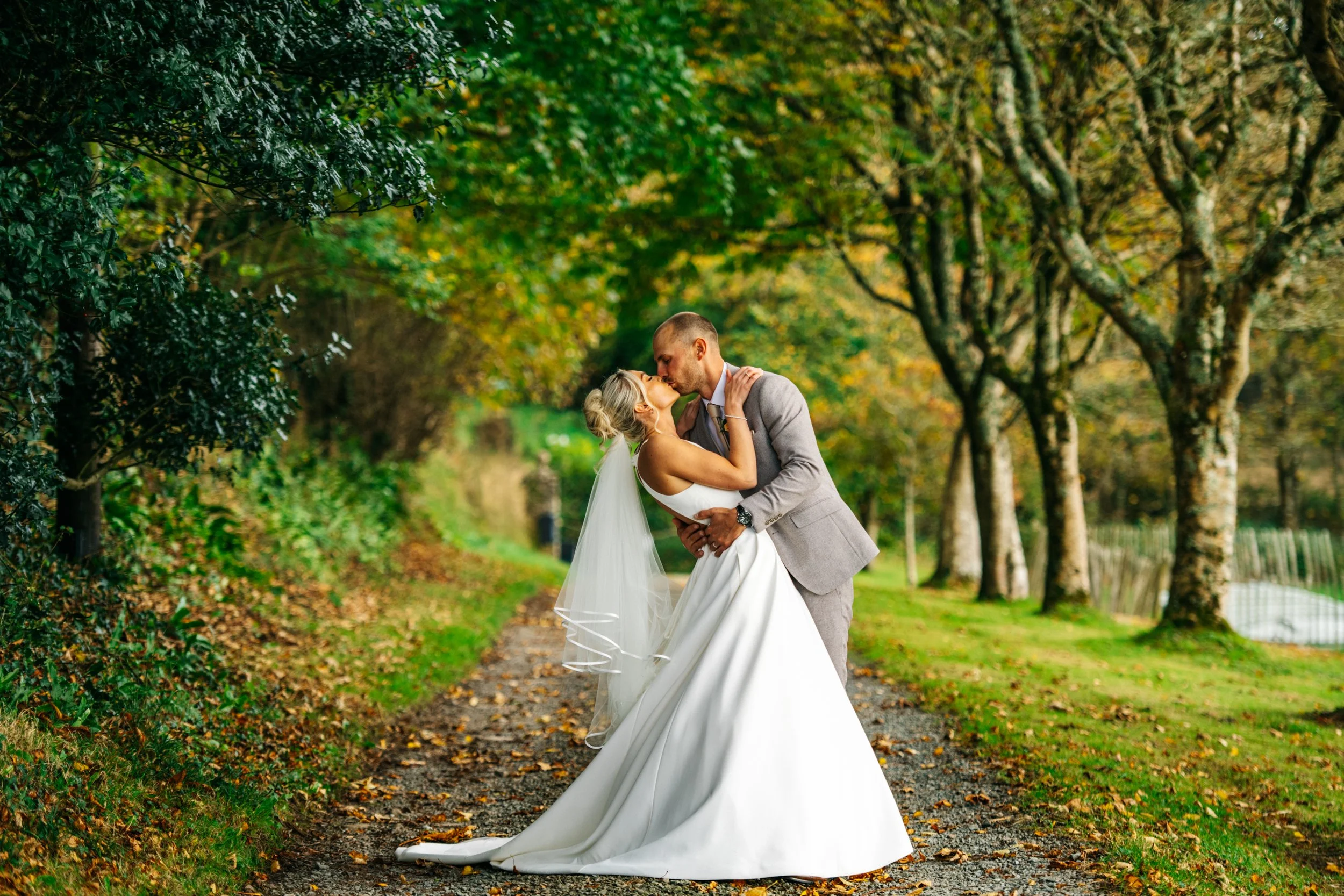 A bride and groom kissing on a tree-lined pathway during their wedding day, surrounded by autumn foliage.