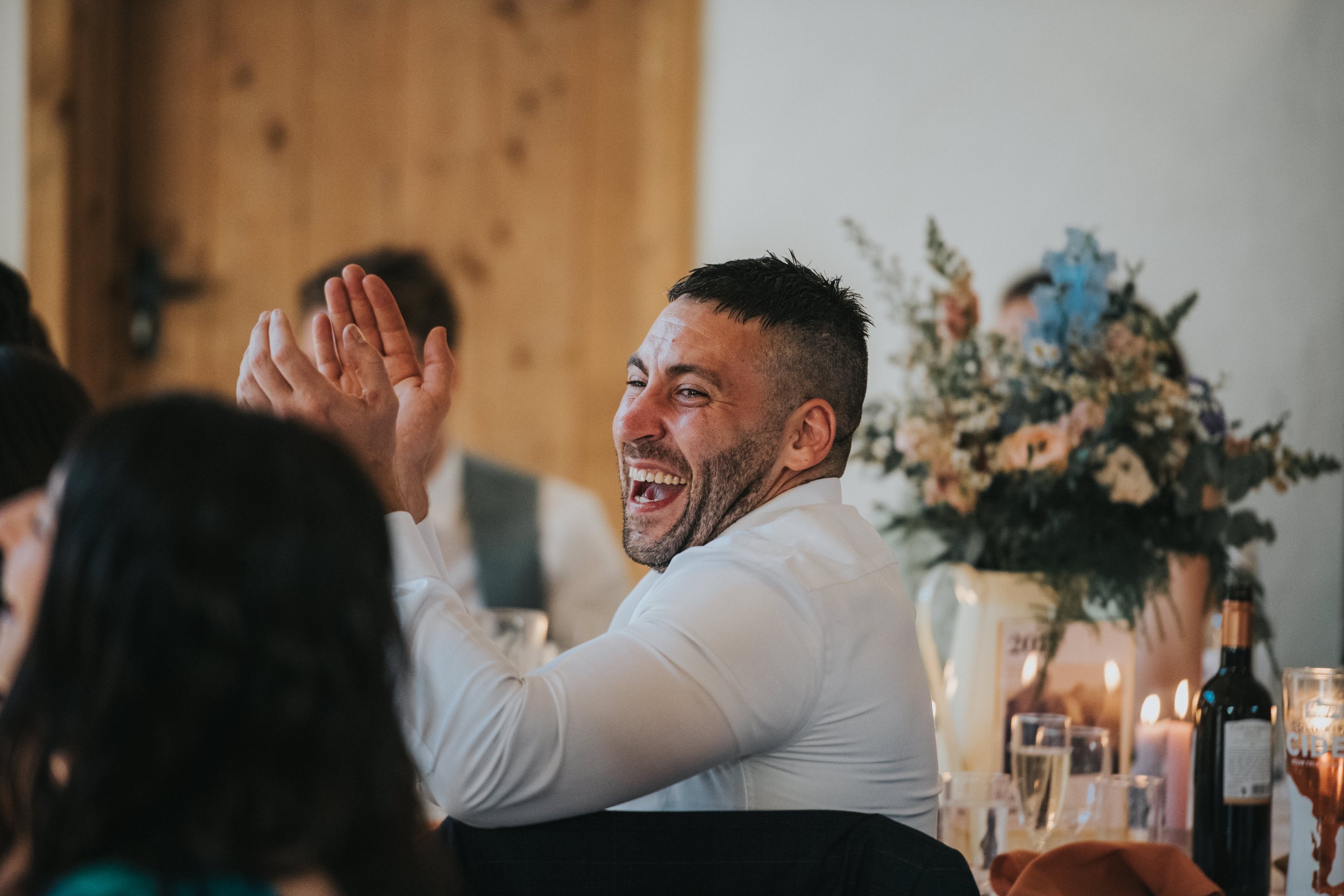 A man in a white shirt laughing and clapping at a celebration, surrounded by other people, with a floral arrangement and candles on the table.