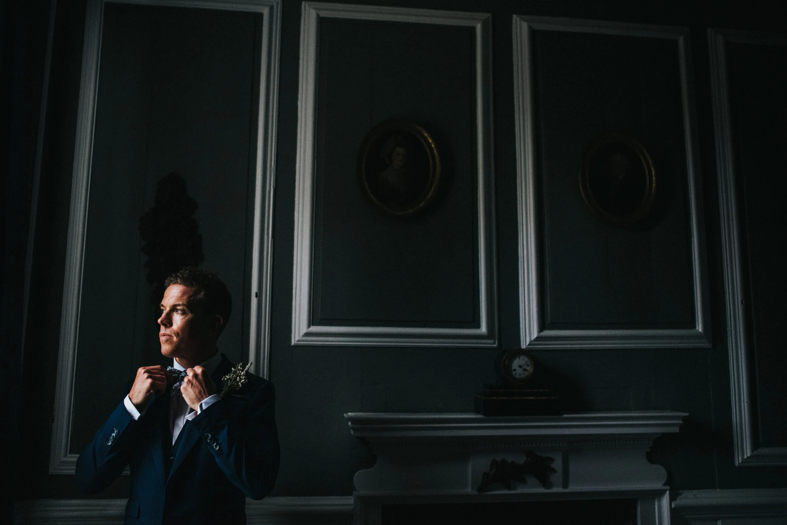 A man dressed in a formal dark suit adjusting his bow tie in a dimly lit room with elegant dark green walls, framed portraits, a clock, and a fireplace.