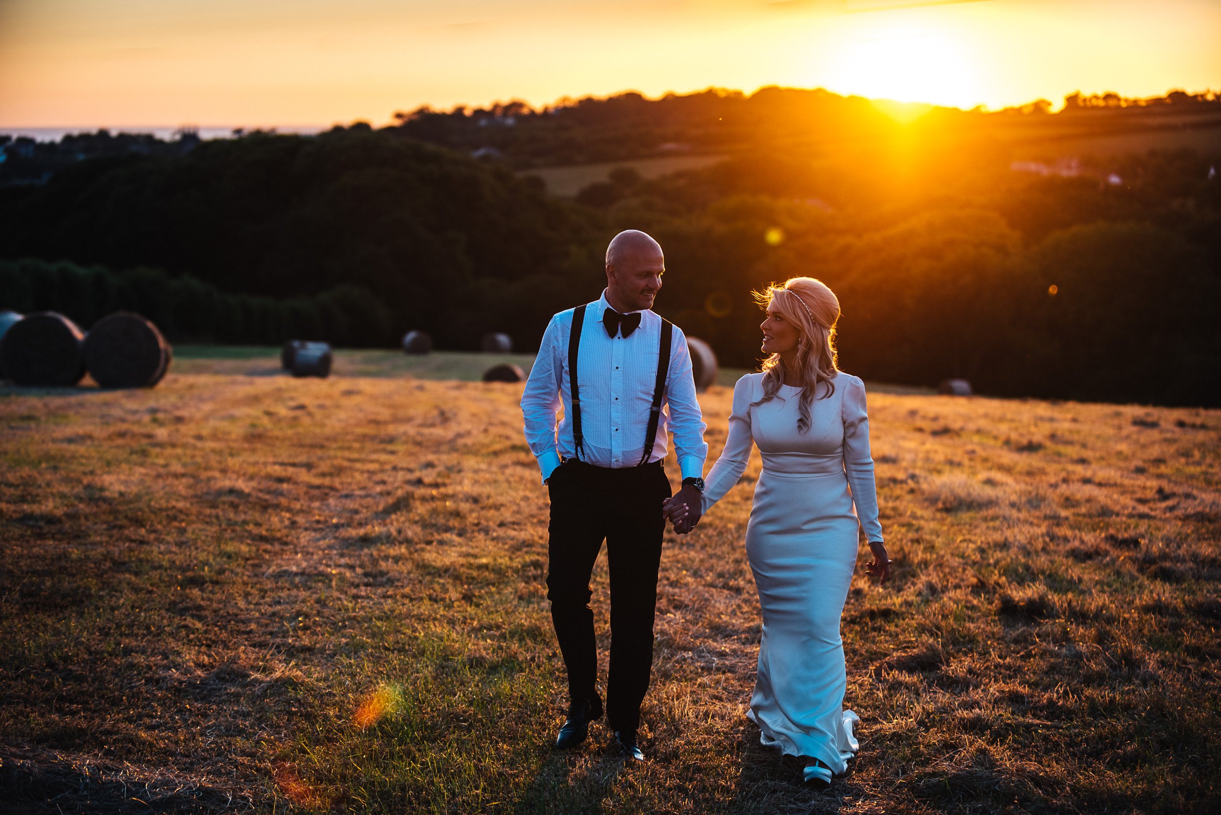 A couple holding hands walks through a field at sunset, with hay bales in the background.