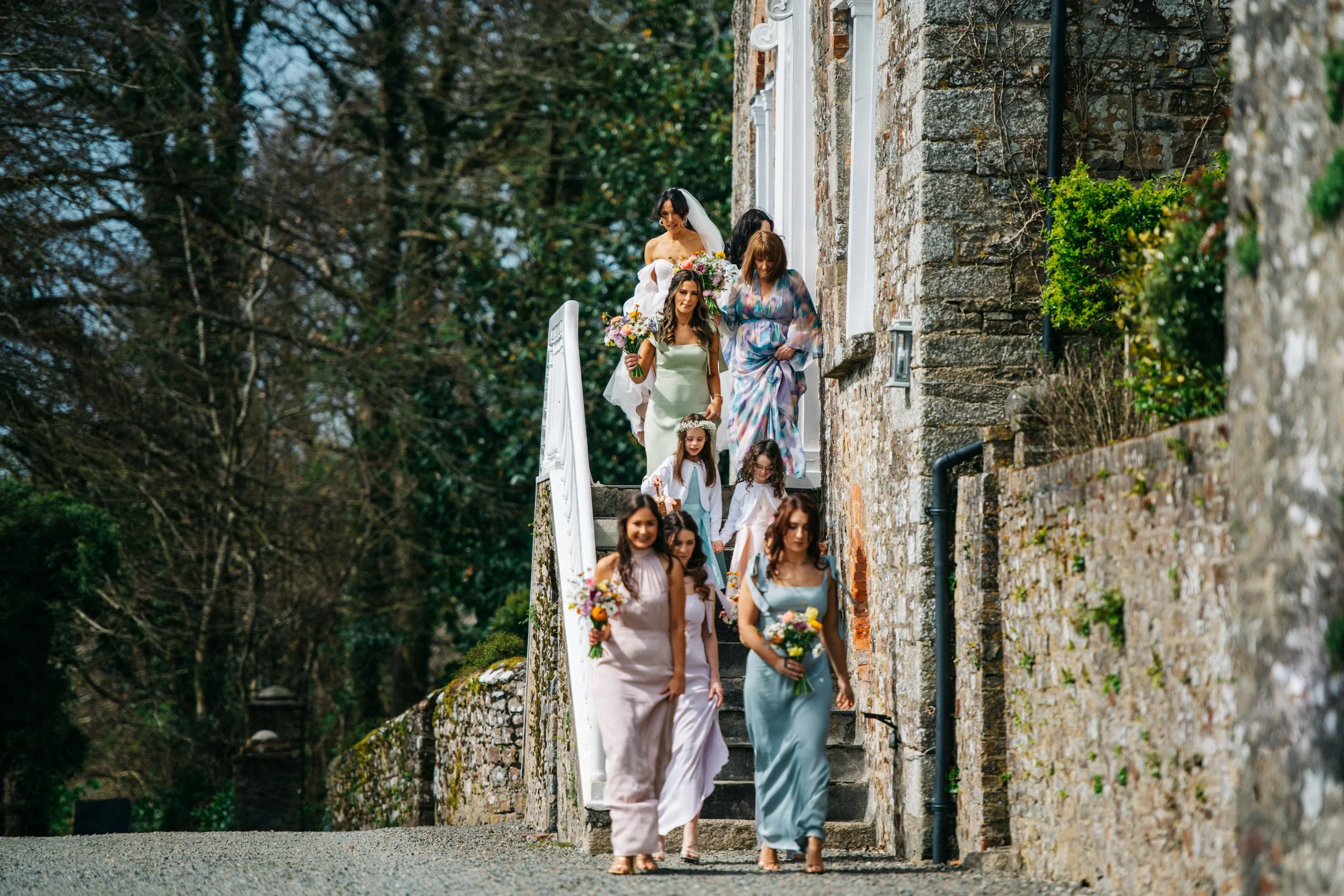 A bride and several women and girls walking down outdoor stone stairs, carrying bouquets, with trees in the background.