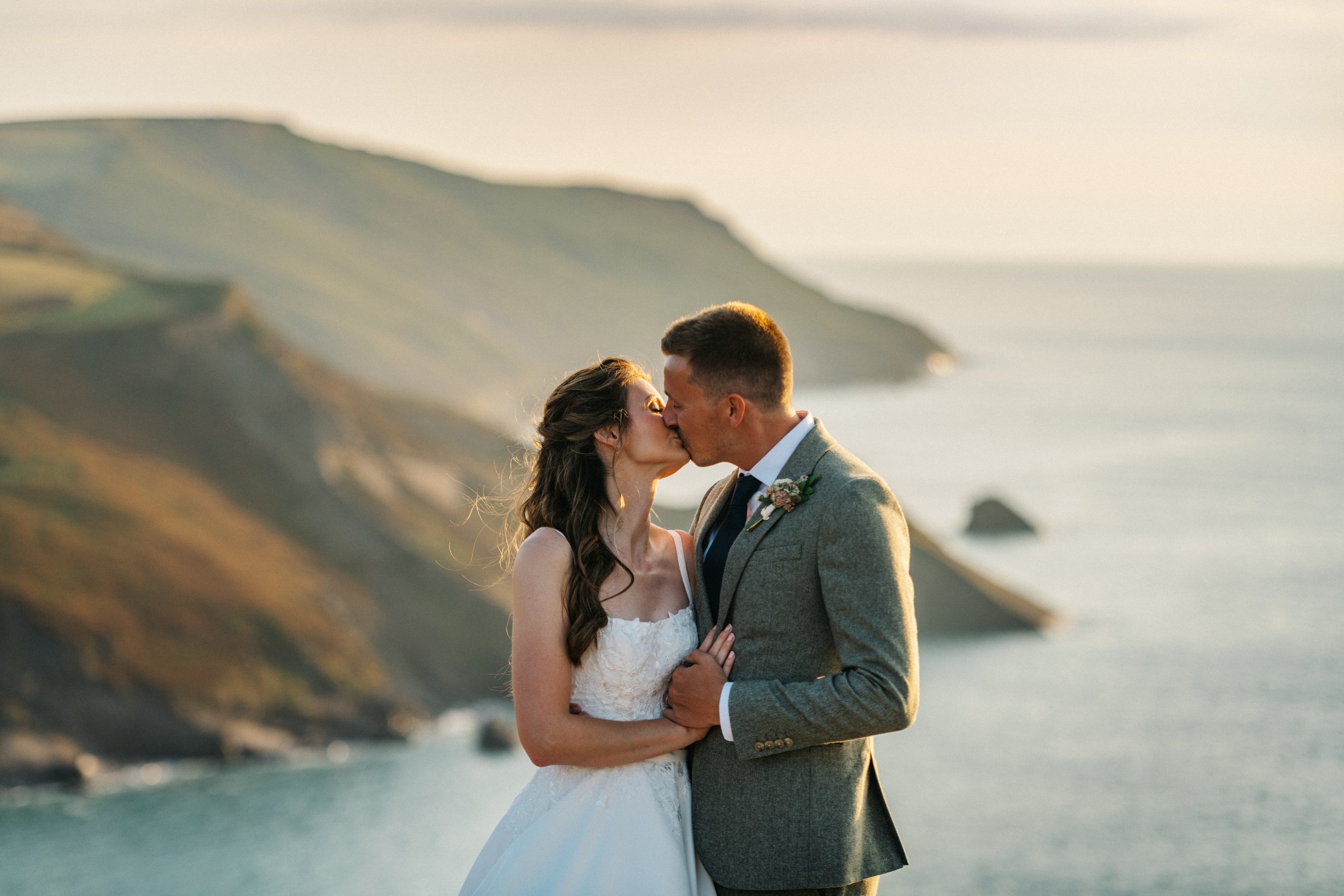 A bride and groom kissing outdoors near the coast with cliffs in the background during sunset.