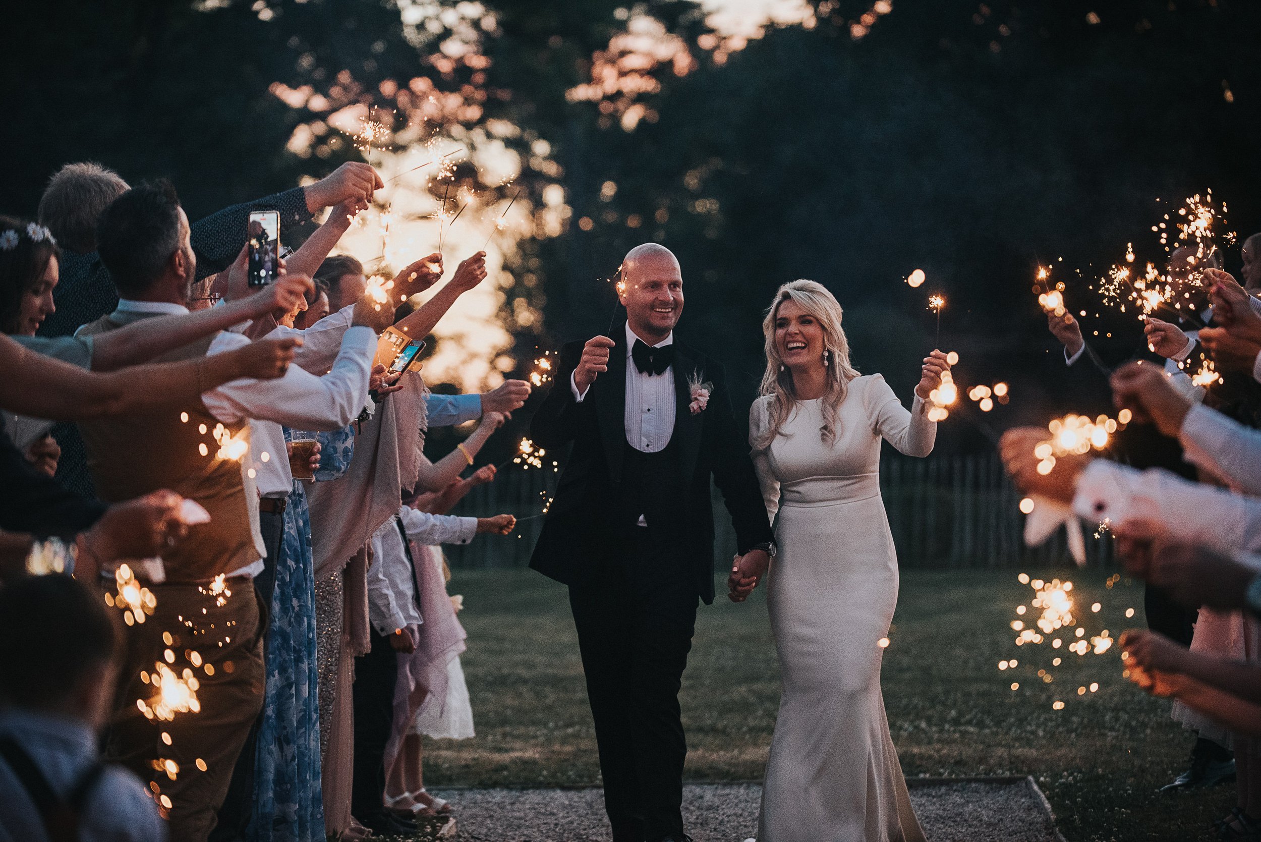 A newlywed couple holding hands and smiling as they walk through a crowd of wedding guests holding sparklers at dusk or evening.