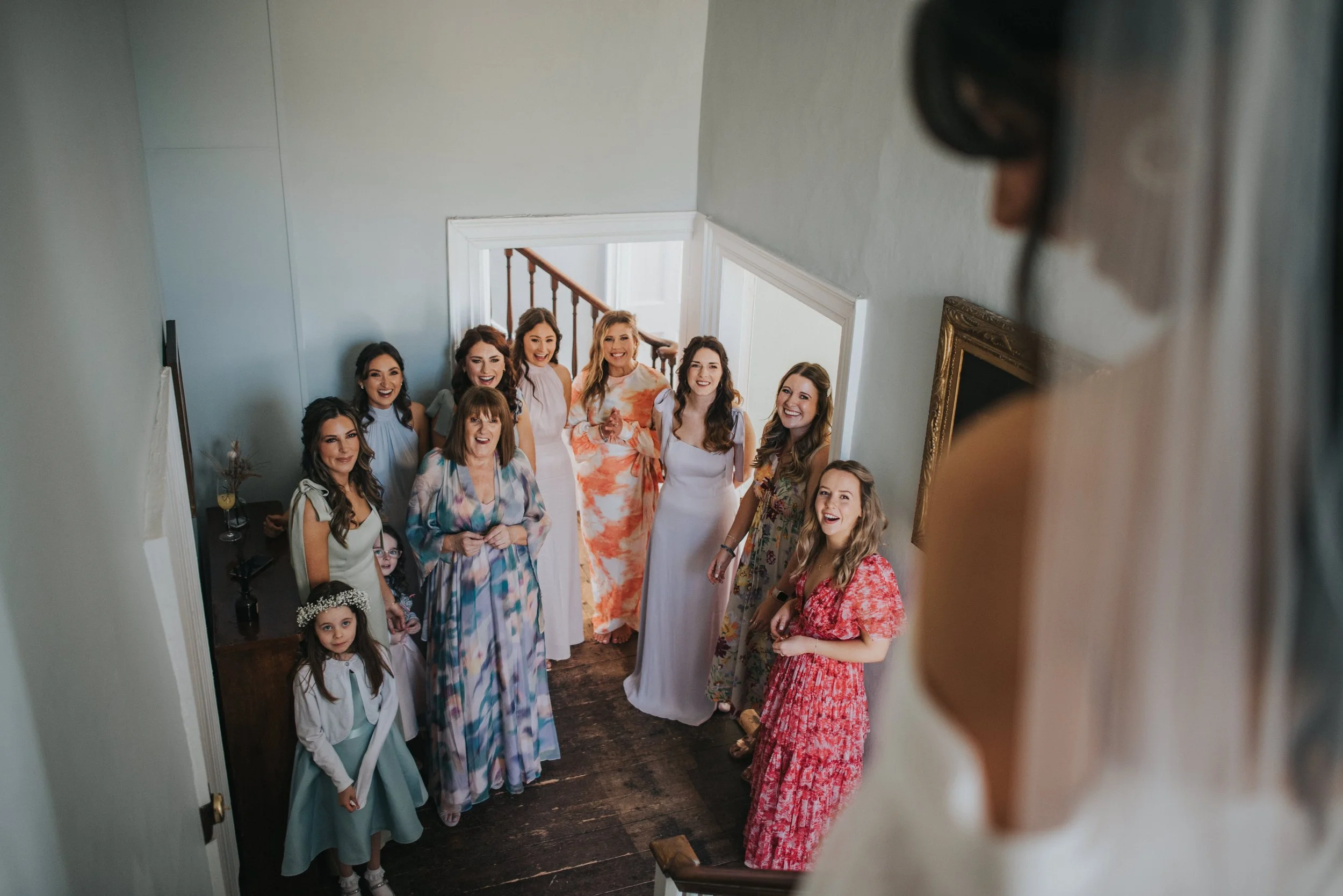 A group of women and girls gathered at the bottom of a staircase indoors, smiling and looking up towards a woman in a white dress, who is partially visible at the top right corner of the image.
