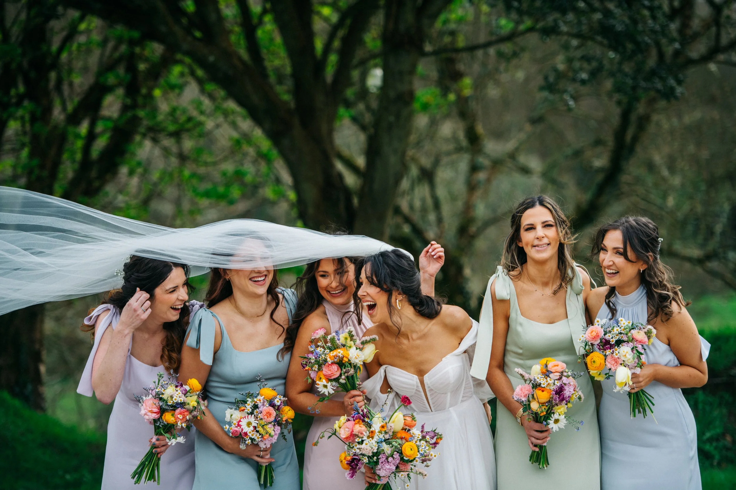 A group of six women at a wedding, standing outdoors in a park with trees in the background. One woman in the center is wearing a white wedding dress holding a bouquet, while the other women are in pastel dresses, holding smaller bouquets, and smilin