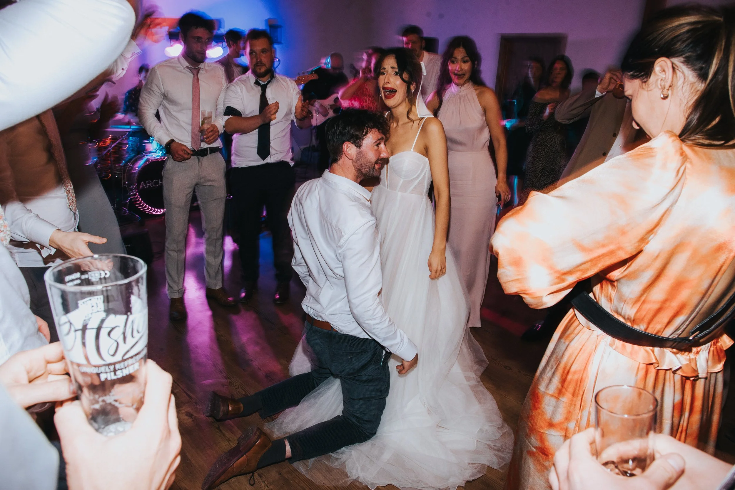 A wedding reception with guests dancing and celebrating. The bride and groom are in the center, with the groom kneeling and the bride smiling. Guests surround them, some holding drinks and others dancing.