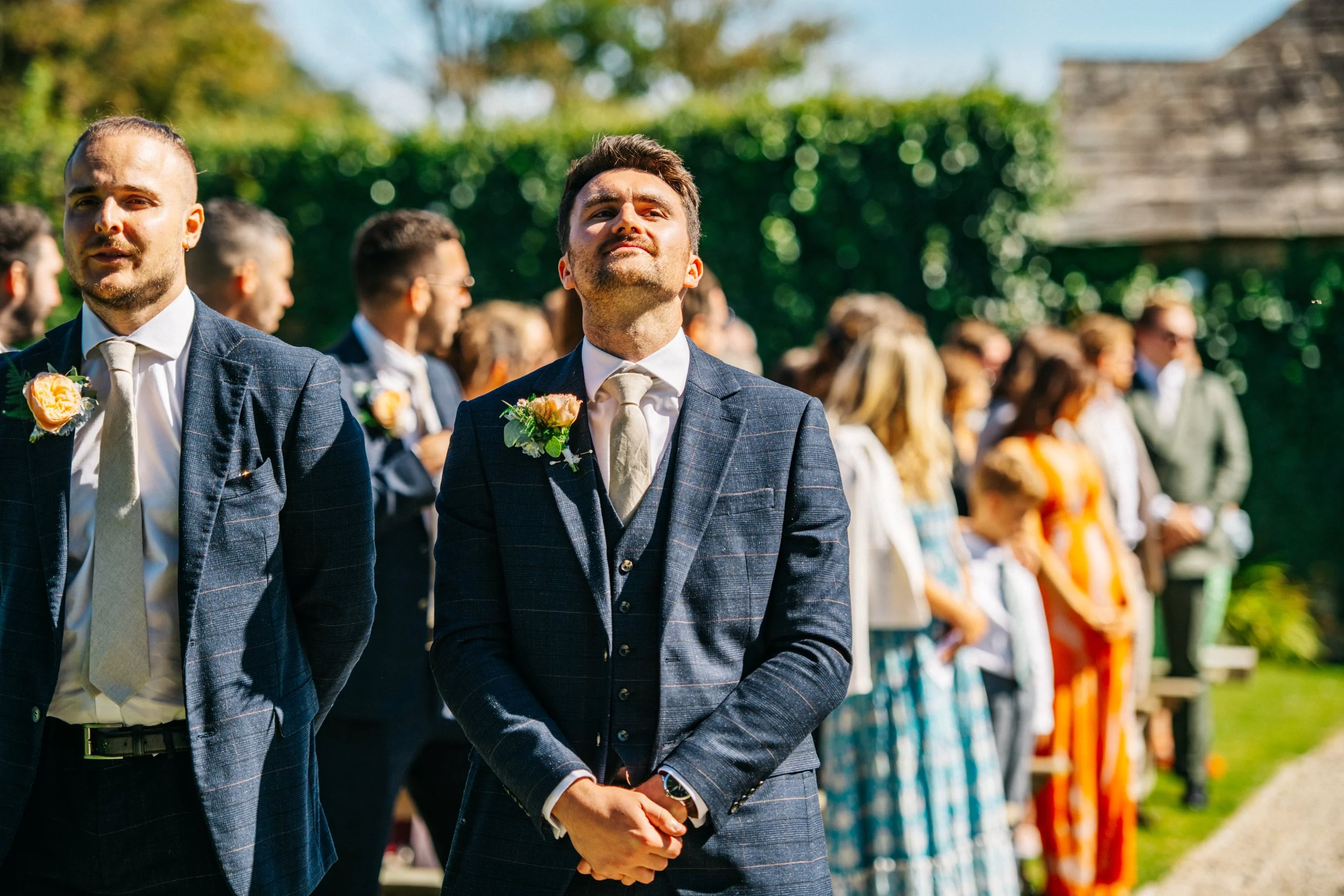 Groom and wedding party during outdoor wedding ceremony in sunlight, people wearing formal attire with floral boutonnieres, standing in line with trees and green hedges in the background.