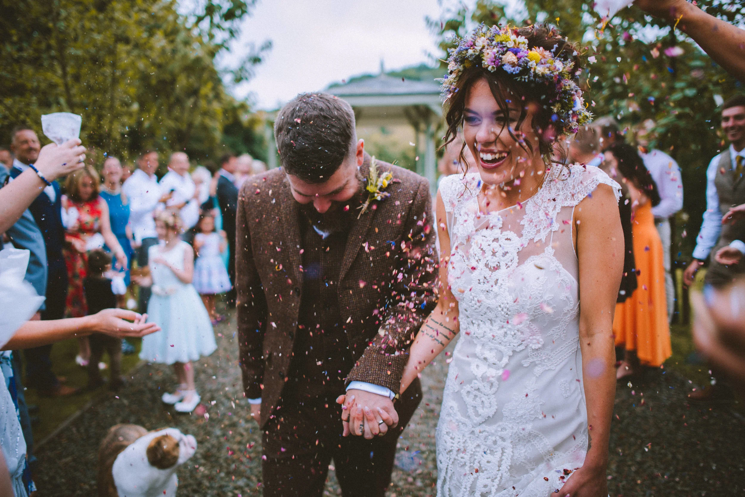 Bride and groom celebrating, holding hands, and smiling after their wedding, surrounded by guests and confetti, outdoors during the day.