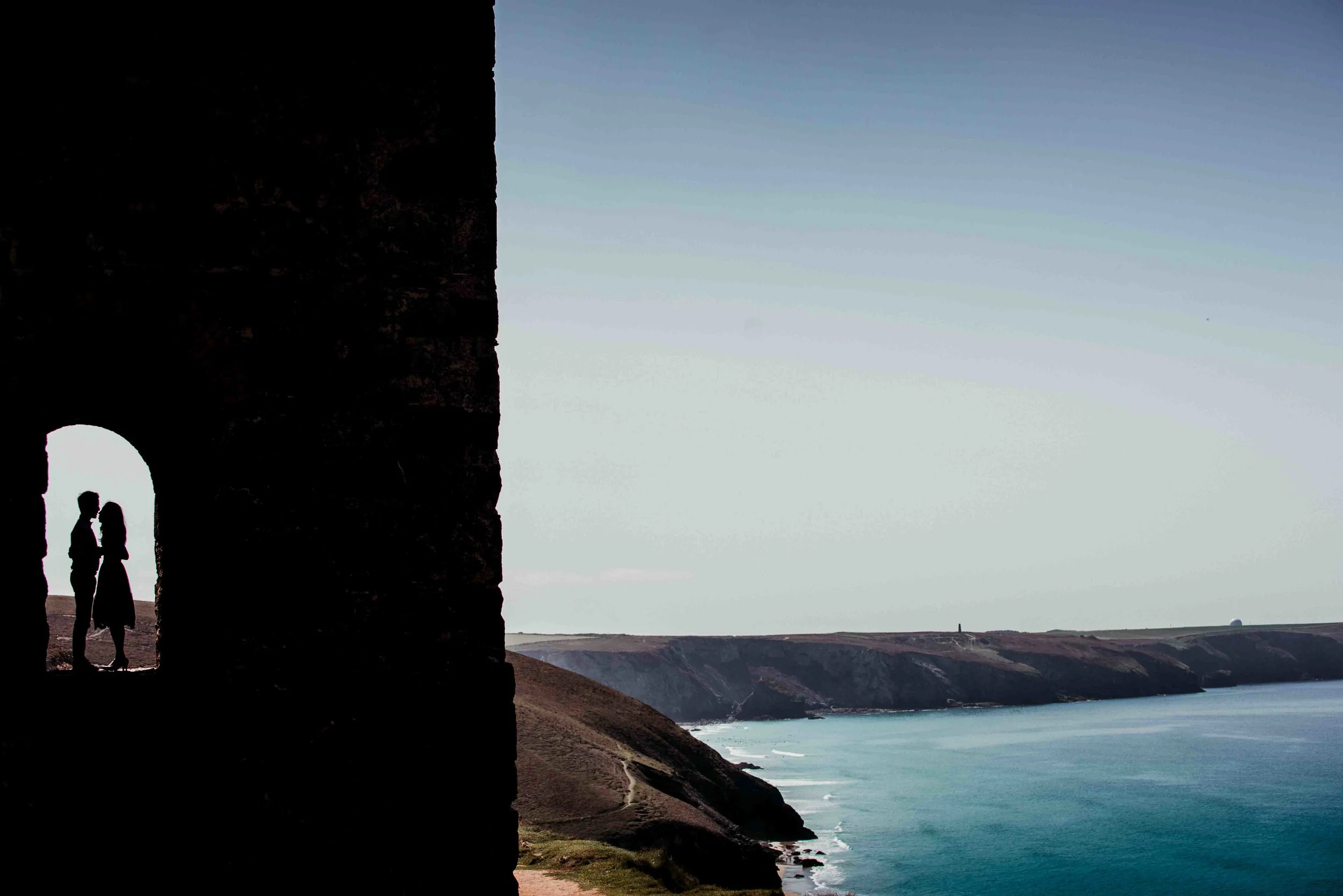 Silhouette of a couple standing under an archway on a cliff overlooking the ocean with a coastline in the background.