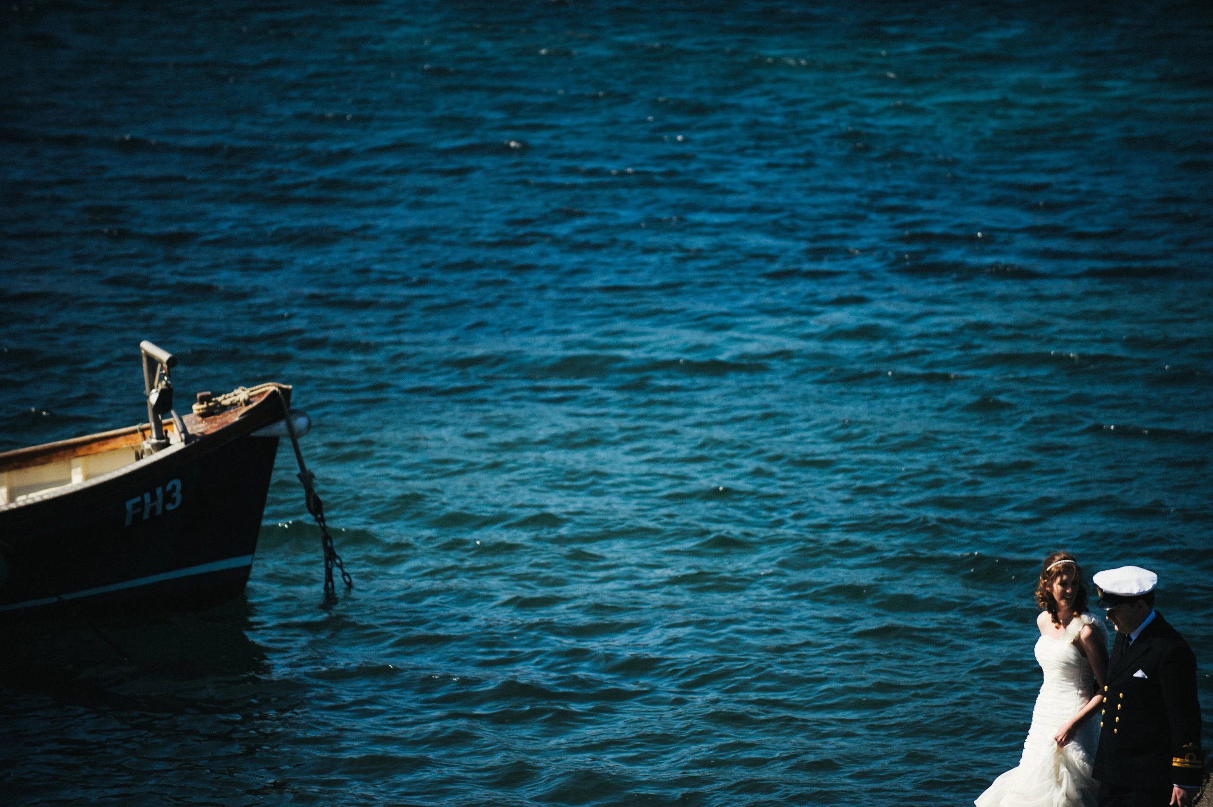 A woman in a white wedding dress and a man in a navy officer uniform walking along the water near a boat in a harbor