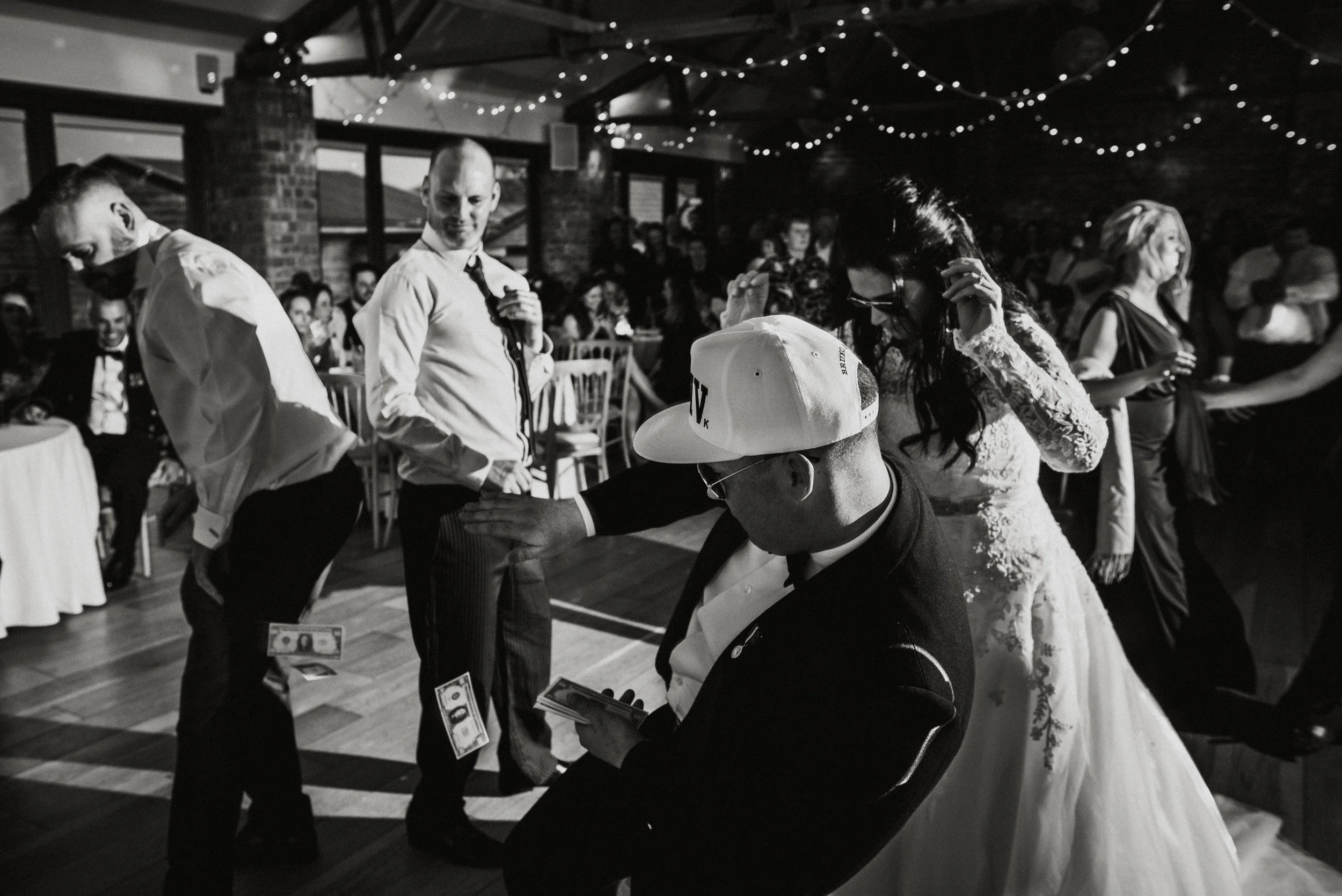 People dancing and socializing at a wedding reception with string lights overhead, a man with bills in midair, and a person wearing a cap with a wedding dress in the background.