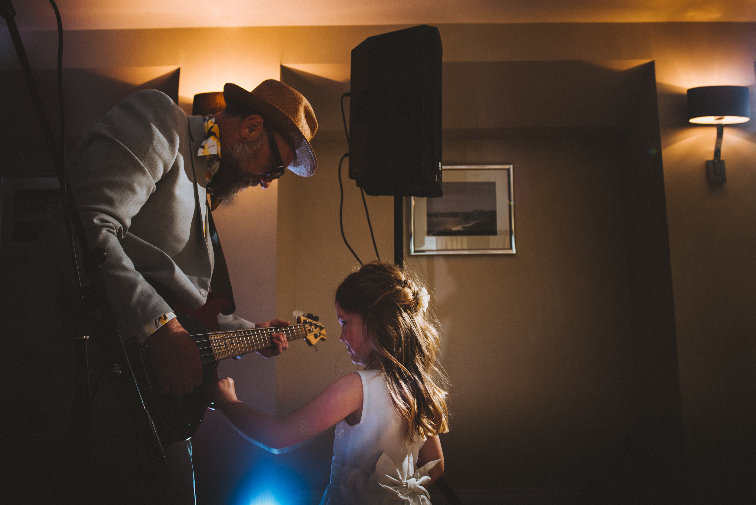 A man playing guitar and a young girl interacting on a dimly lit stage or room with soft lighting and framed pictures on the wall.