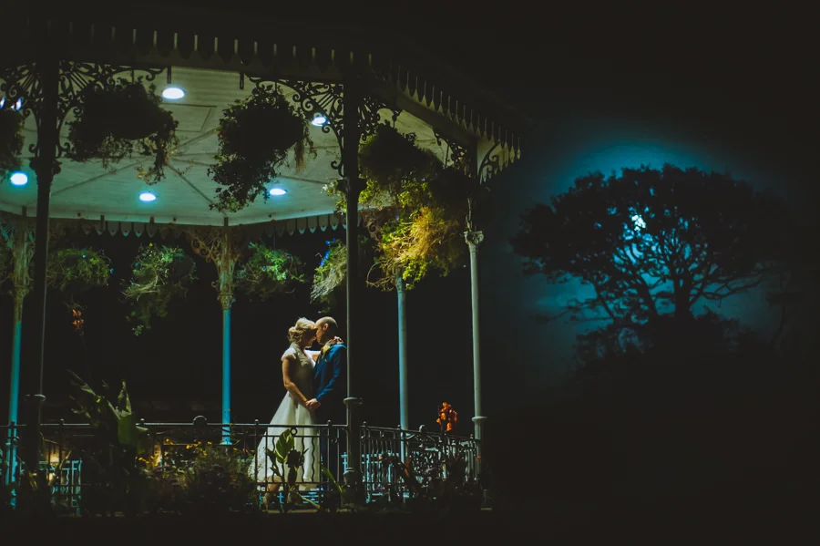 A couple dressed in wedding attire sharing an intimate moment under a decorated gazebo at night, with a moonlit tree silhouette in the background.