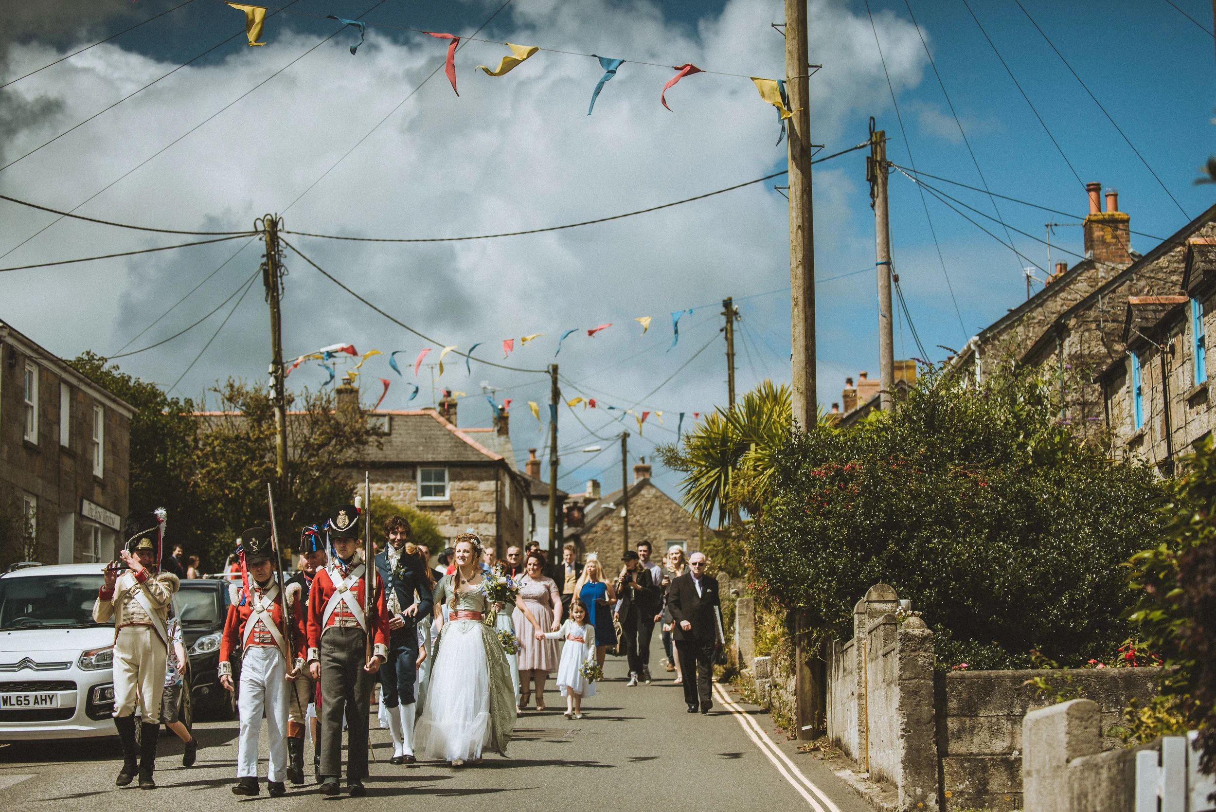 A wedding procession walking down a small street decorated with colorful bunting on a partly cloudy day.