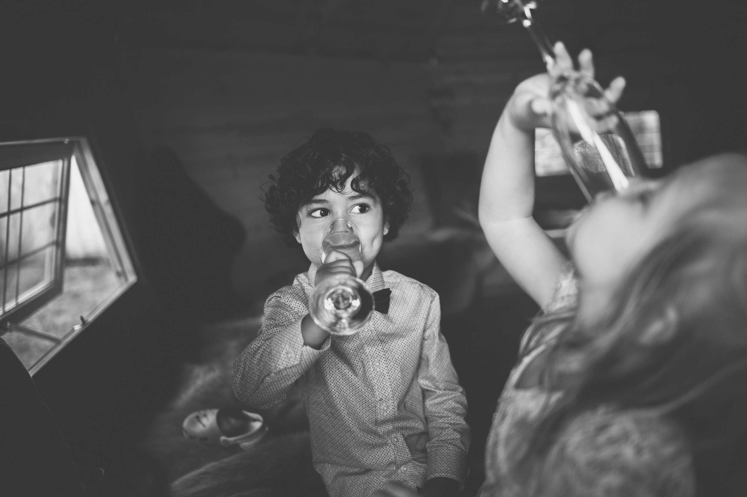 A young boy with curly hair and a bow tie drinking from a glass, while a girl with long hair is holding an glass up to her mouth in a room with a wooden ceiling.
