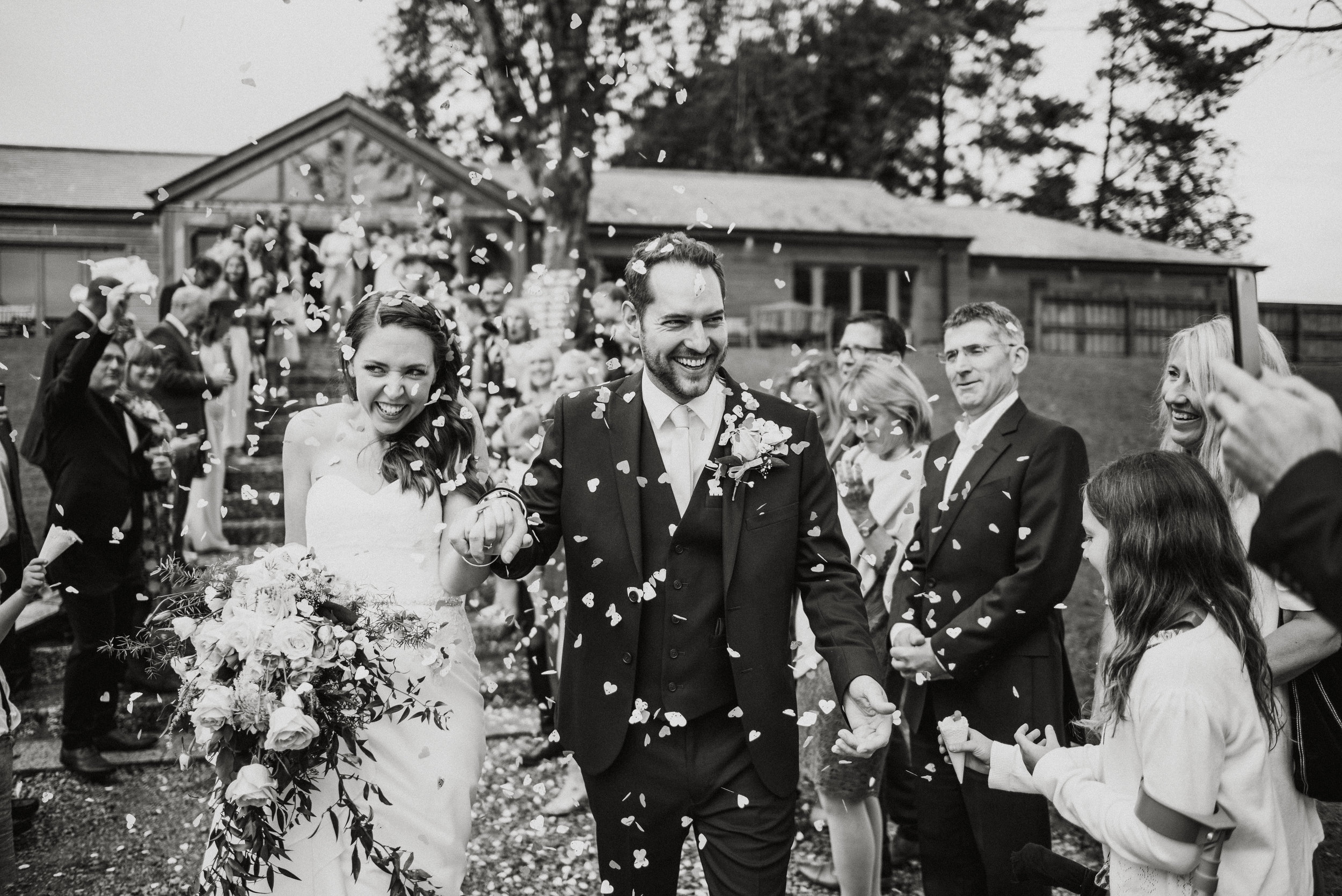 Black and white photo of a wedding celebration with a newlywed couple walking hand-in-hand through a crowd, confetti falling around them, and guests smiling and cheering.