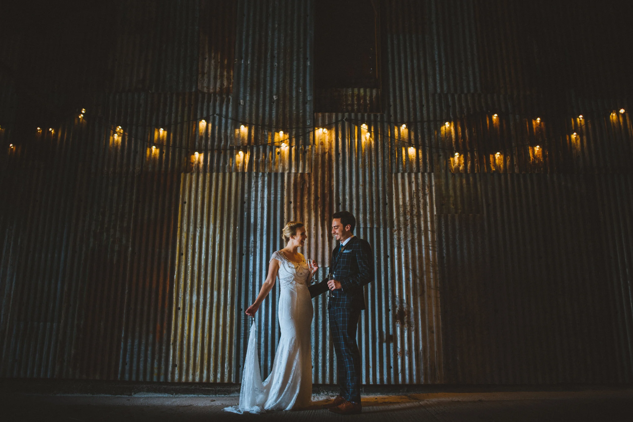 A man and woman in wedding attire stand together, smiling, in front of a rustic, corrugated metal wall with warm string lights overhead at night.
