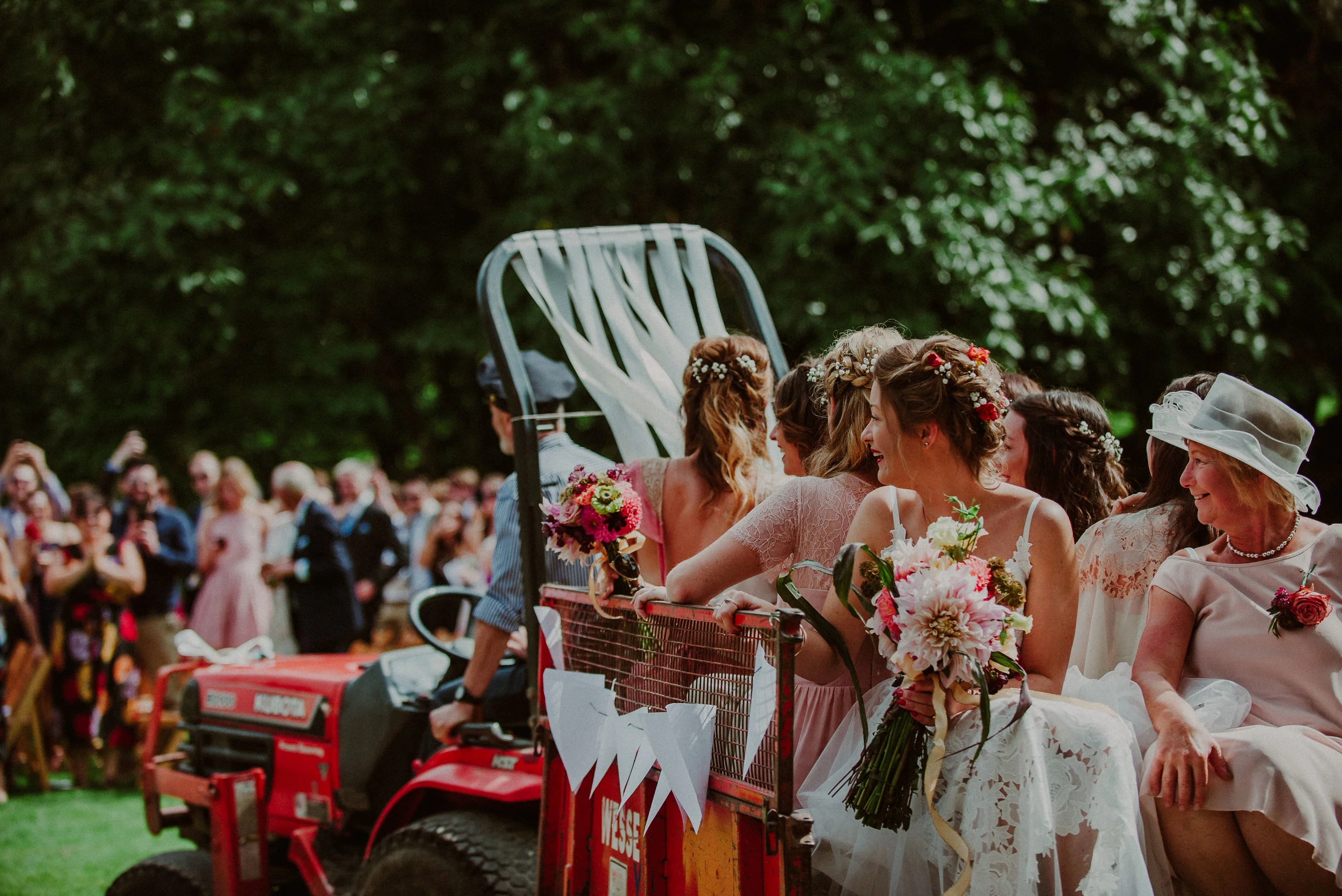 Group of women in wedding dresses sitting on a small tractor decorated with flowers and white ribbons, with wedding guests in the background at an outdoor event.