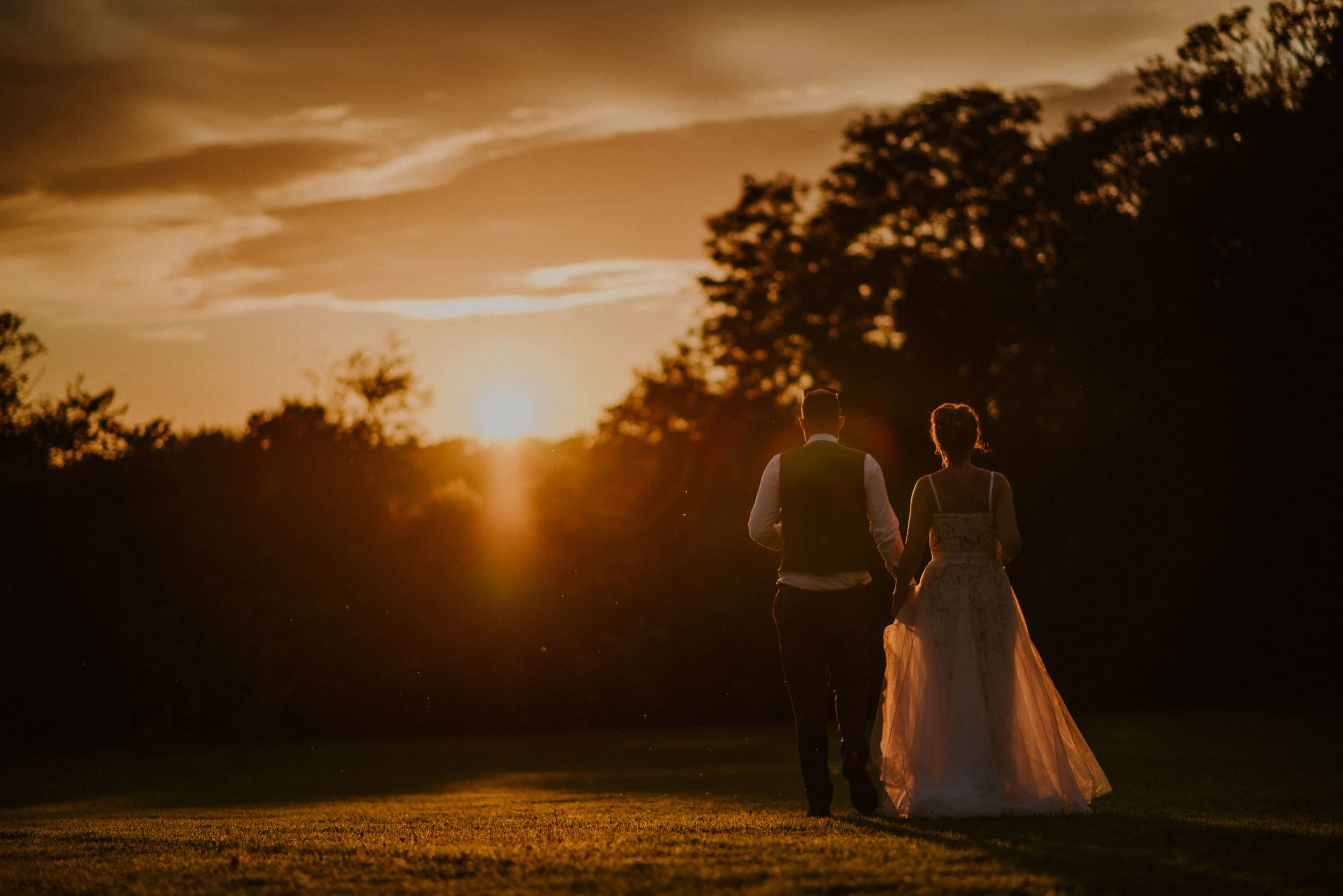 A newlywed couple holding hands walking away in a field at sunset with trees in the background.