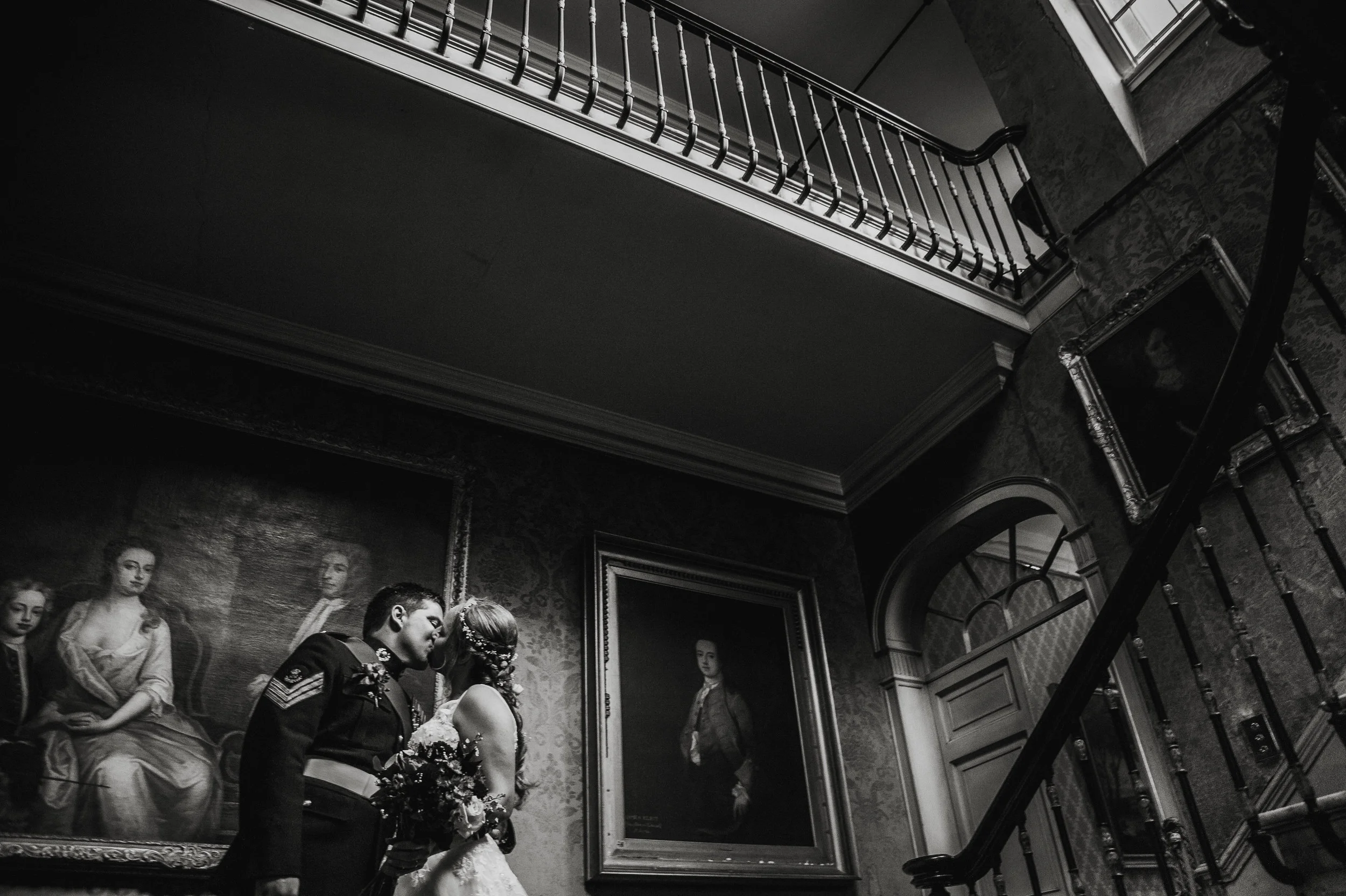 A bride and groom in wedding attire standing close and nose to nose in a dimly lit, elegant room with Victorian-style portraits on the walls.