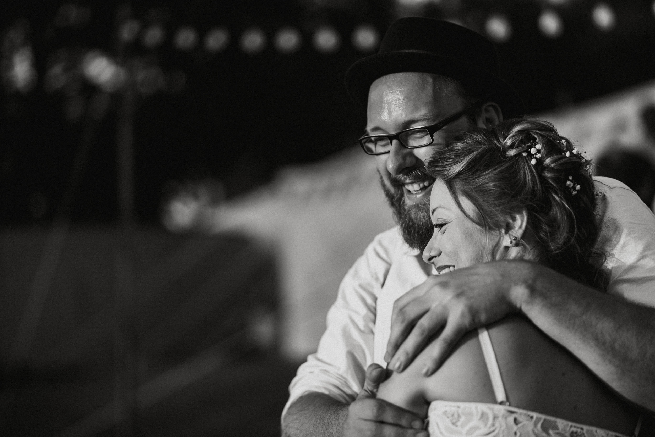A joyful couple sharing a hug, the man wearing glasses and a hat, the woman with styled hair and floral accessories, in a black-and-white photo.