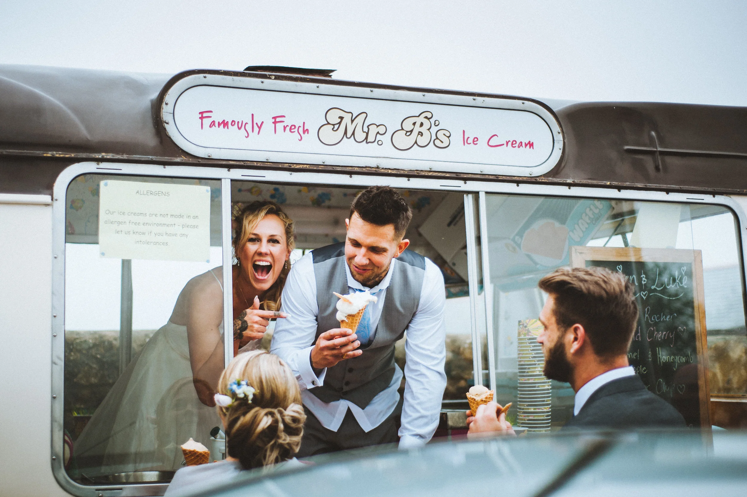 A group of people enjoying ice cream inside a small ice cream truck called Mr. Bs Ice Cream, with a sign saying 'Famycly Fresh Mr. Bs Ice Cream'. They are smiling and interacting, with a woman pointing and a man holding an ice cream cone.